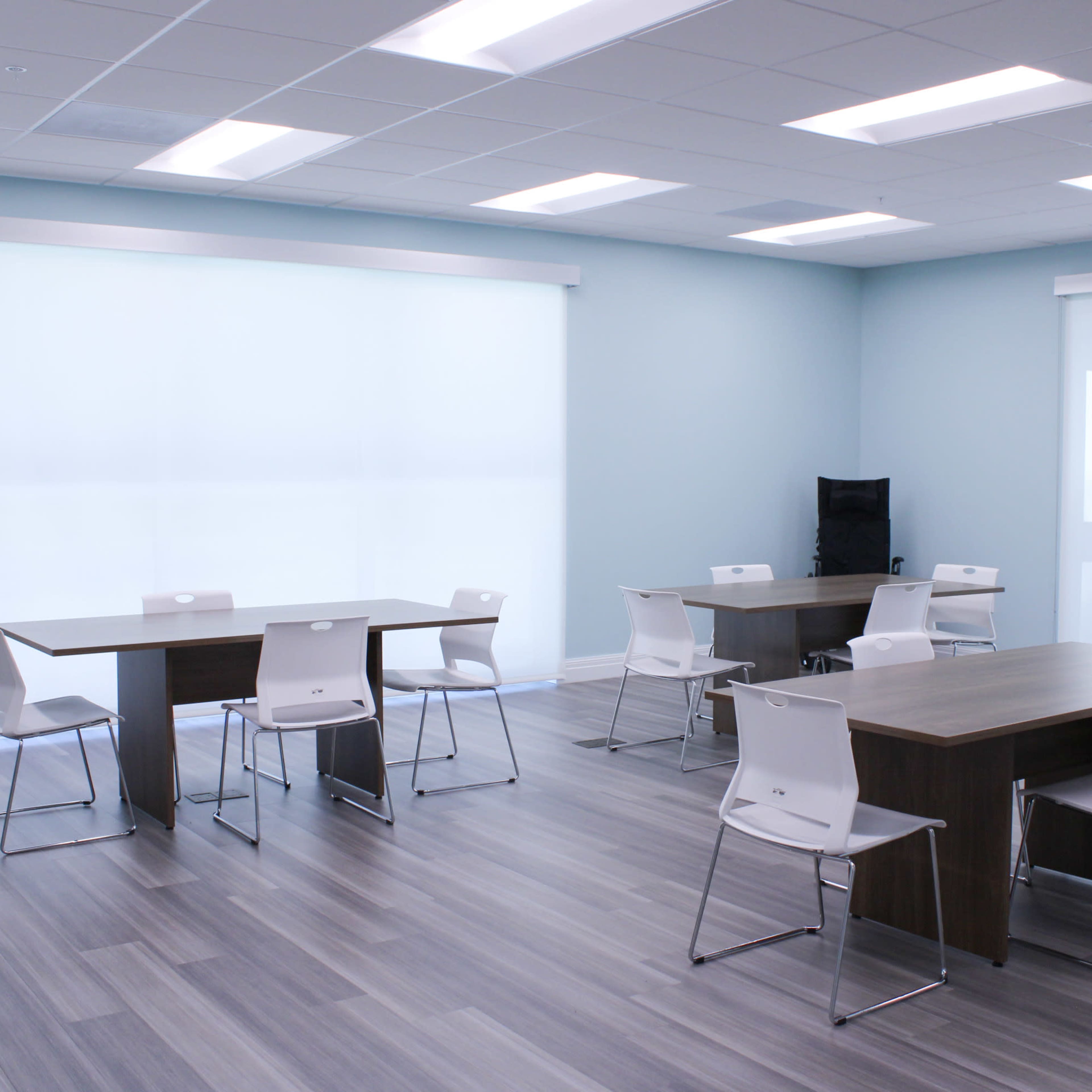 A modern classroom with several tables and chairs arranged on a wooden floor, illuminated by natural light from large windows.