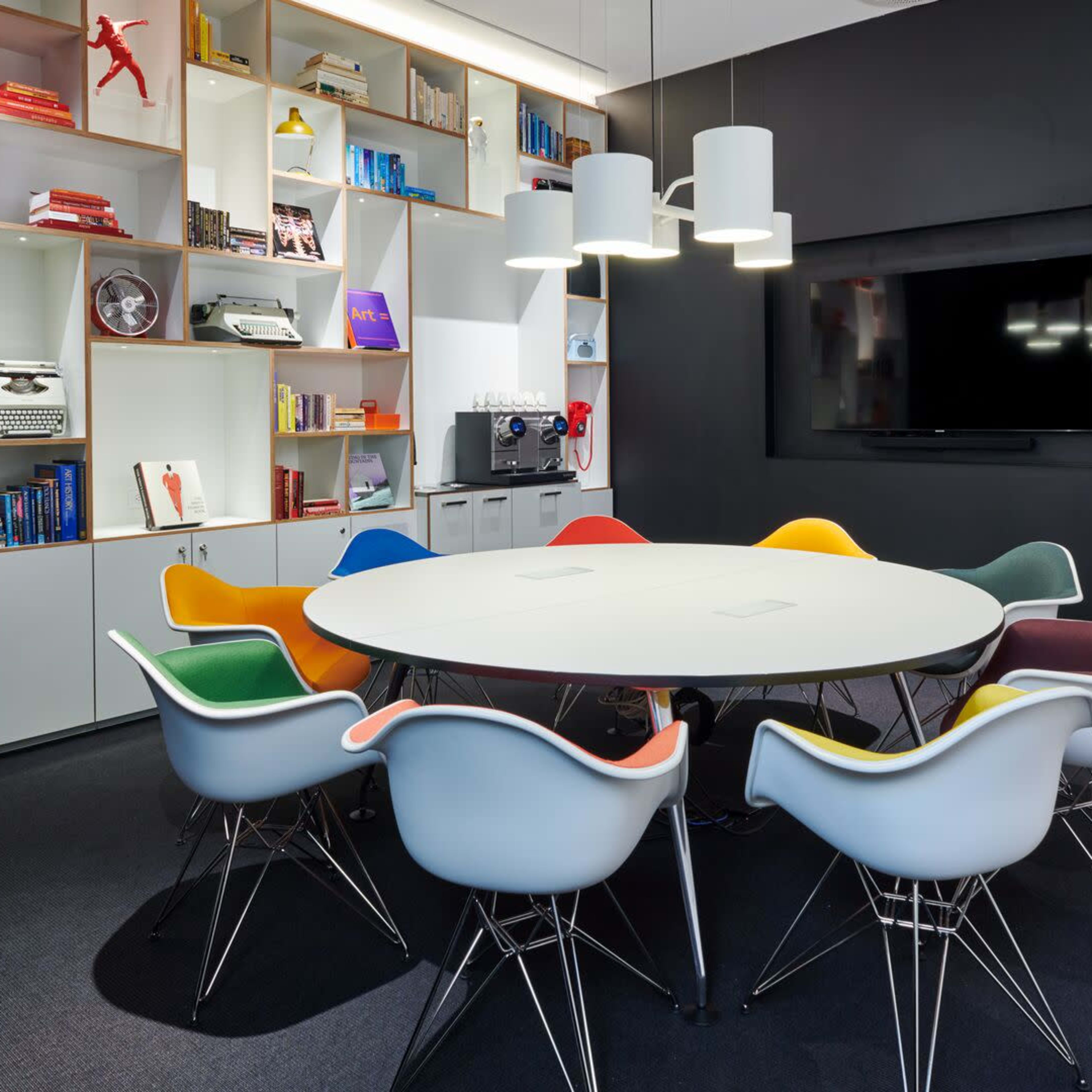A modern conference room features a round table surrounded by colorful chairs, with shelves of books and decorative items in the background.