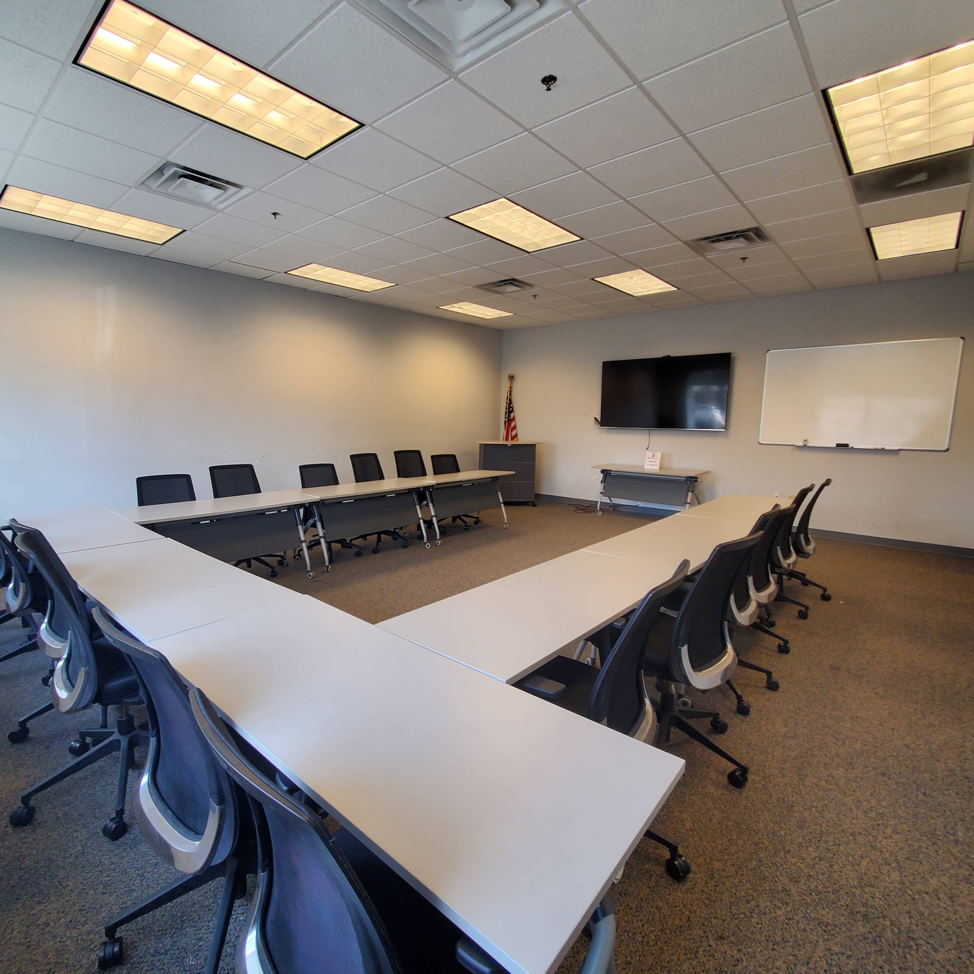 A conference room with a large table arranged in a U-shape, surrounded by black chairs, a whiteboard, and a monitor on the wall.