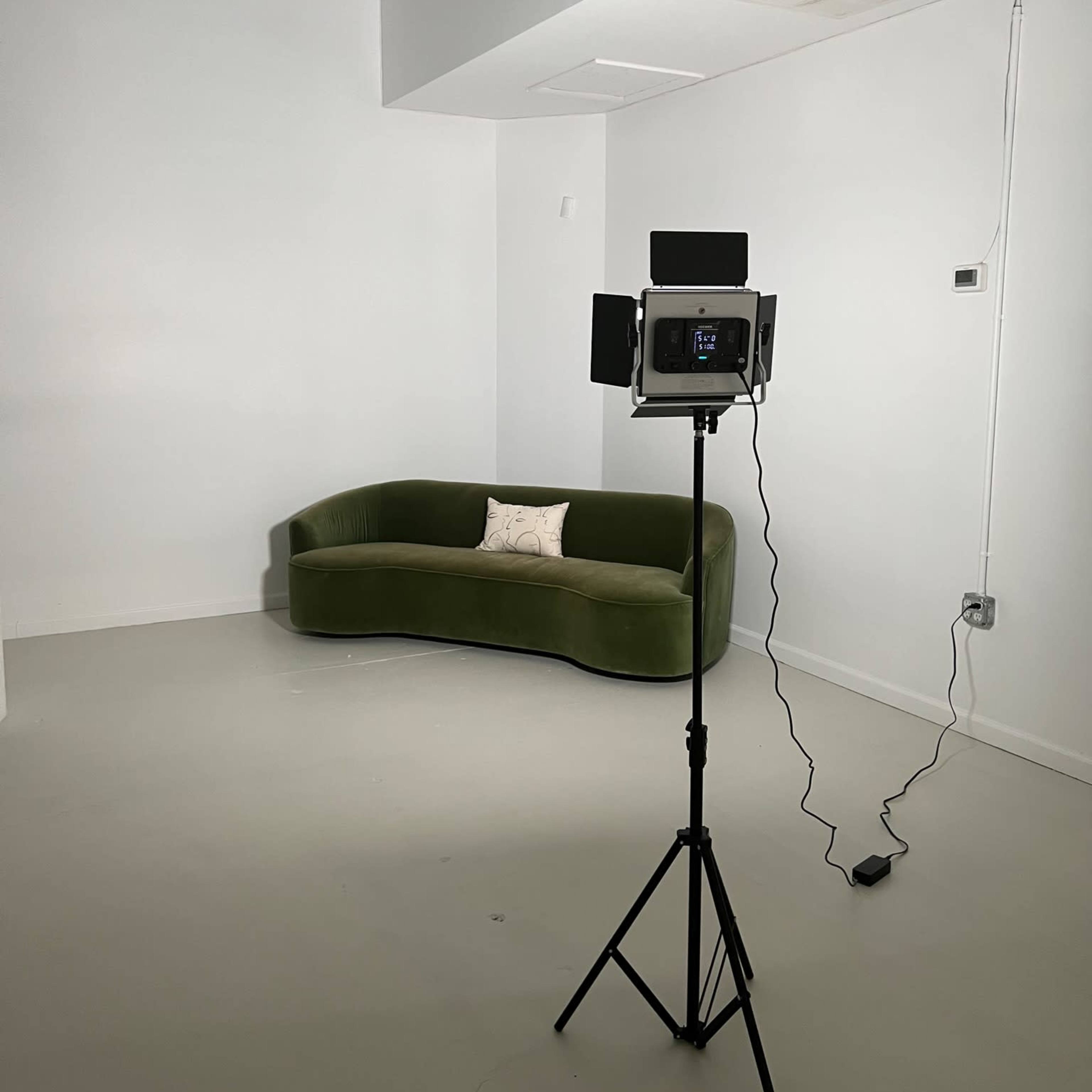A tripod-mounted light stands in front of a green couch in a minimalistic, white-walled room.
