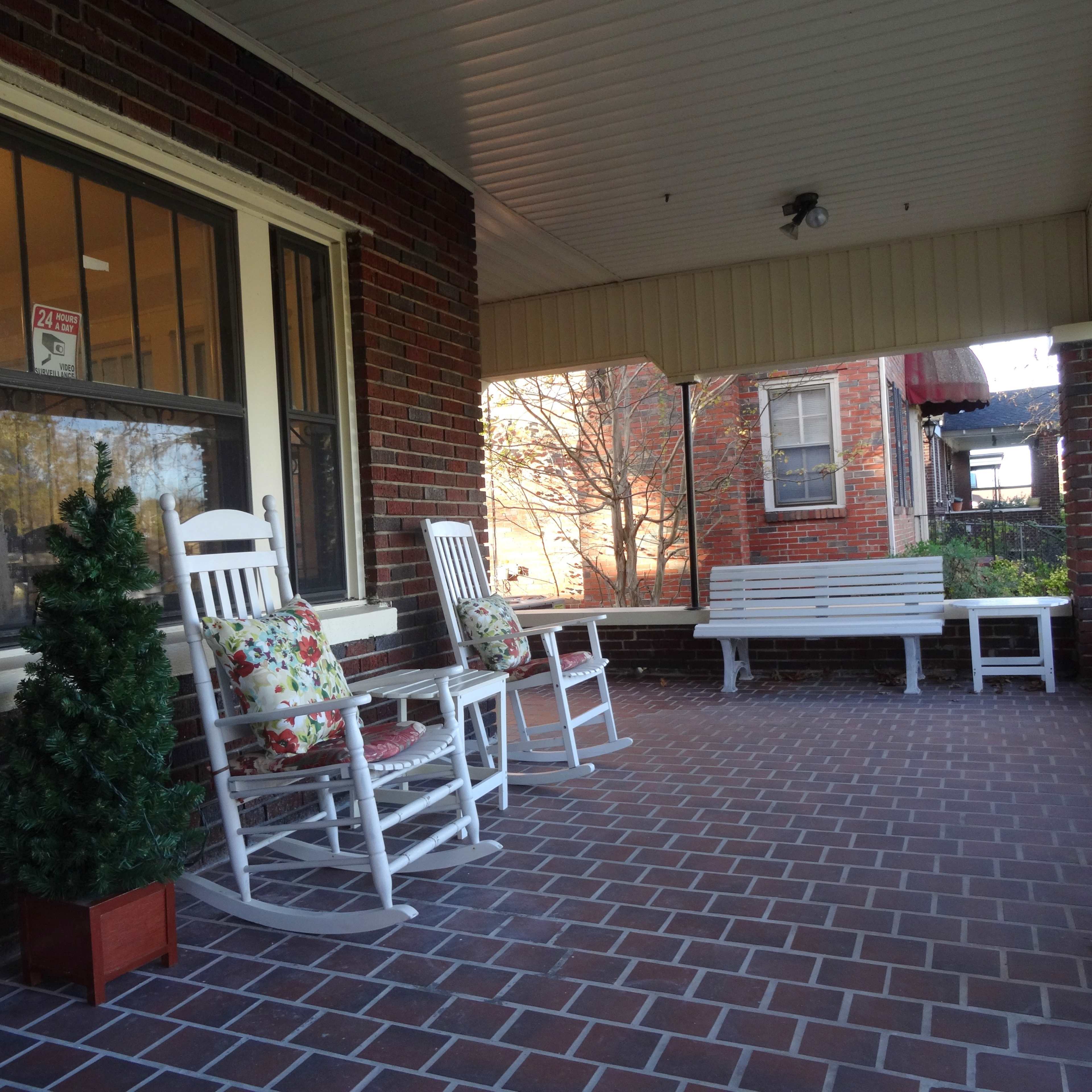 A porch area with two white rocking chairs, a small evergreen plant in a pot, and a white bench, all set against a backdrop of brick walls.