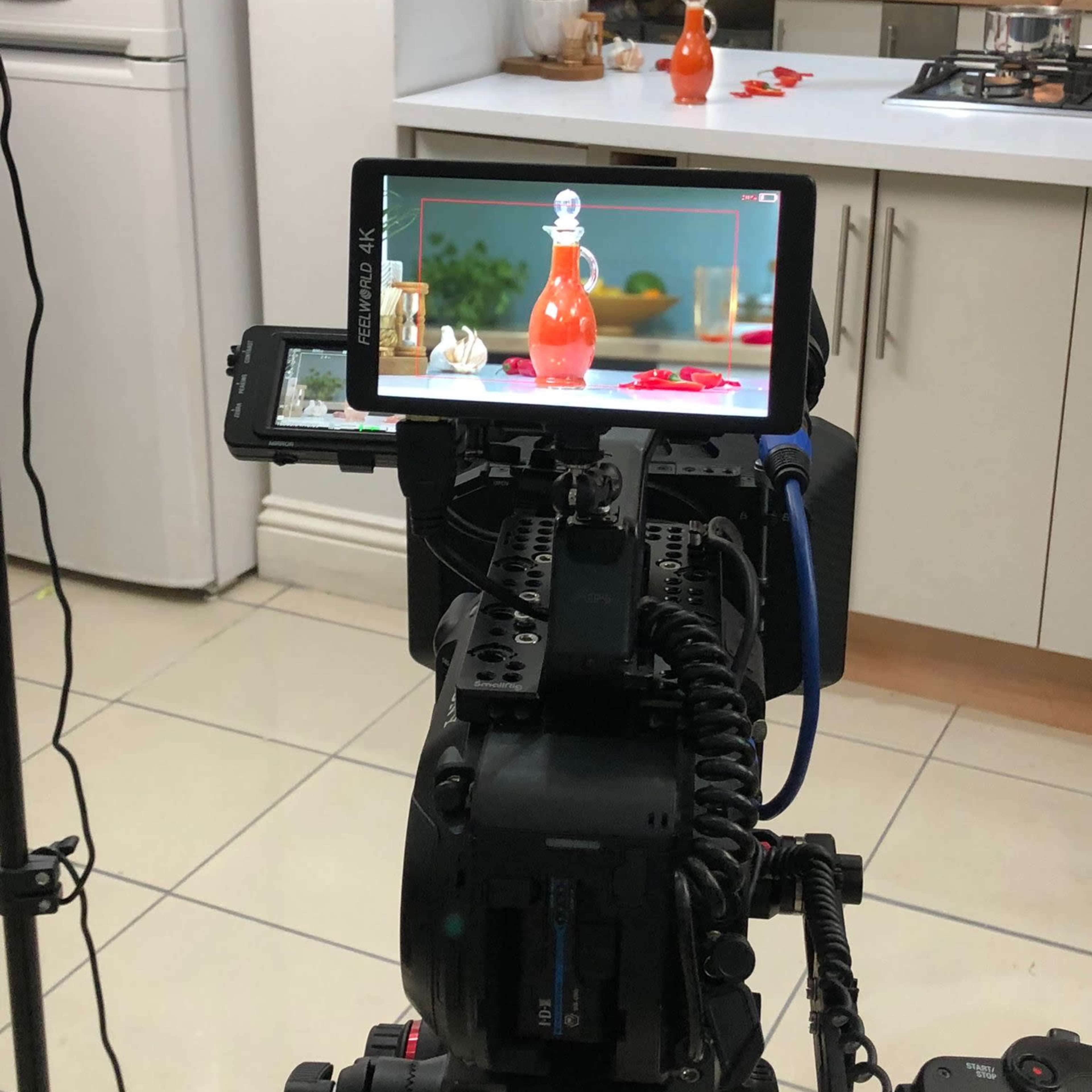 A camera setup on a tripod is focused on a bottle in a kitchen scene with various kitchen items and lighting in the background.