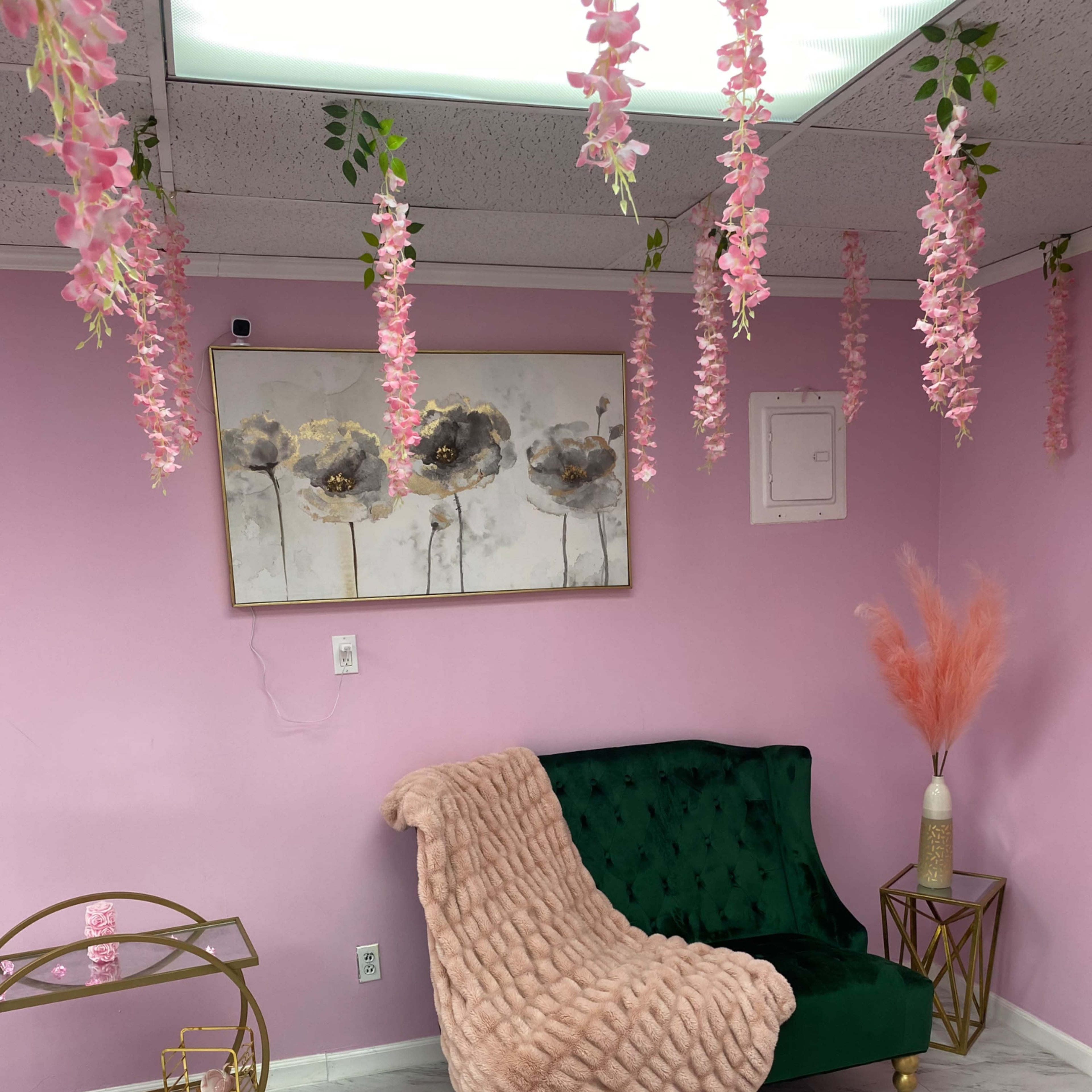 A cozy room with a green velvet loveseat draped with a pink blanket, surrounded by pink floral decorations hanging from the ceiling and a small gold cart in a corner.