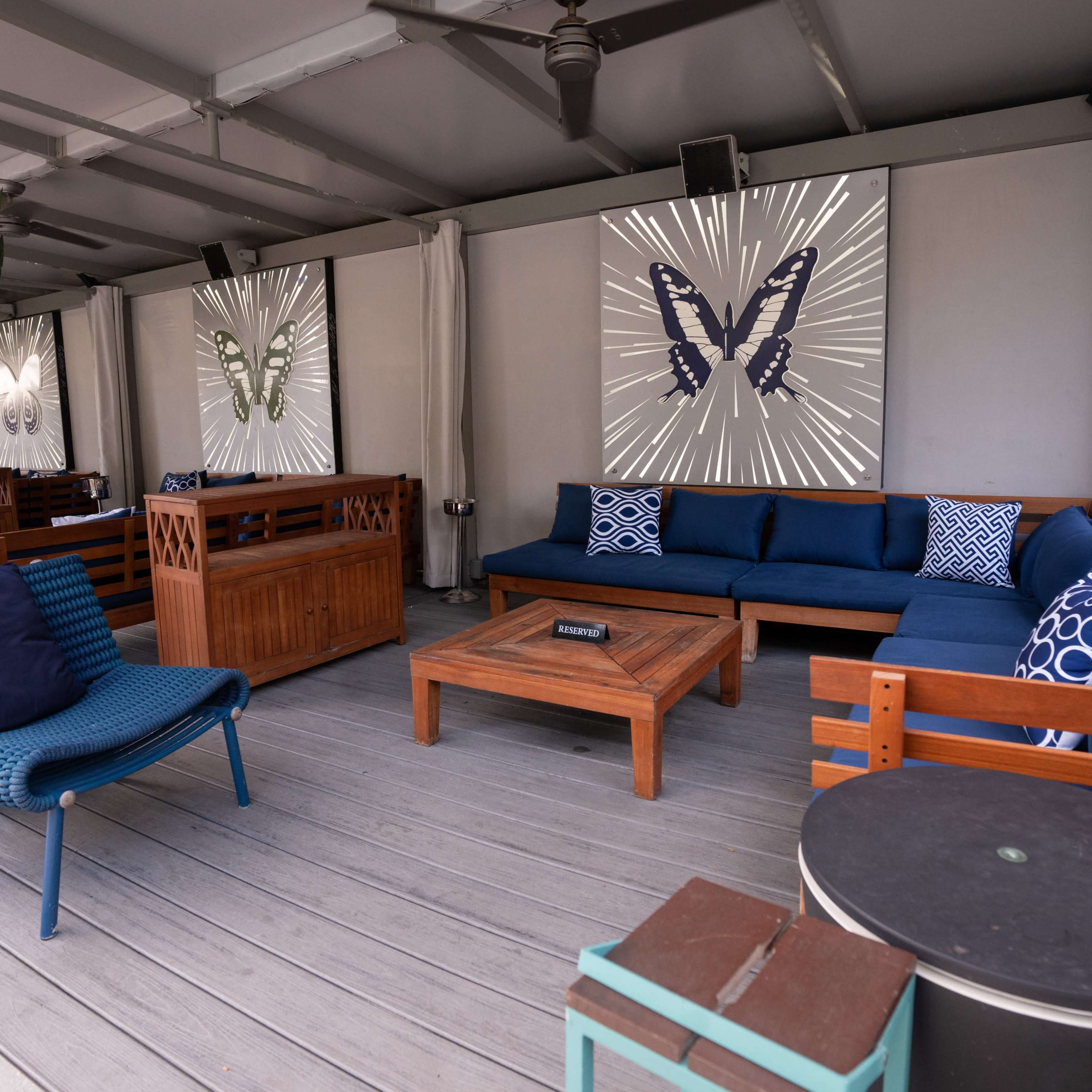 A modern outdoor lounge area with wooden furniture, blue cushions, and two butterfly-themed artworks on the walls.