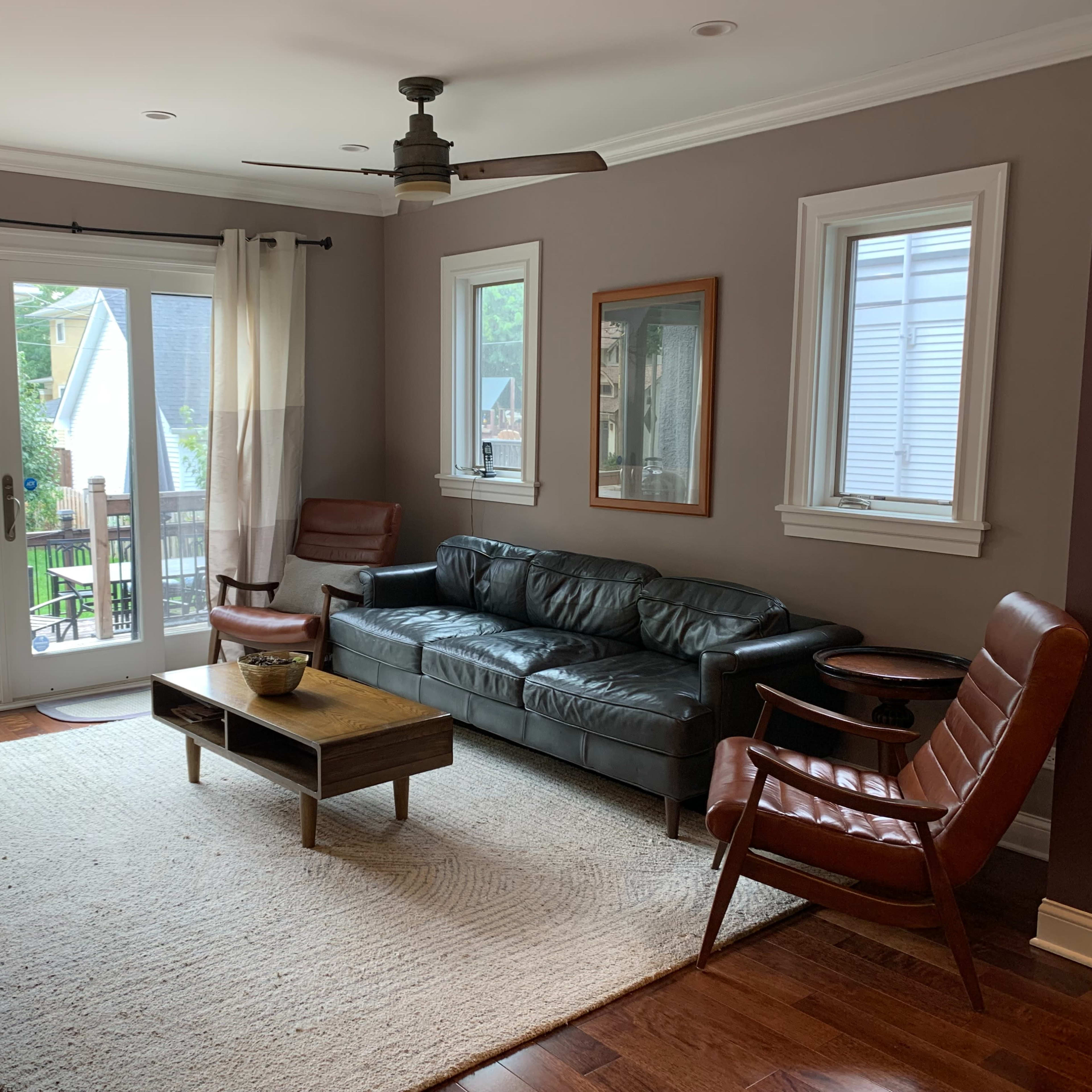 A living room with a dark leather sofa, two brown wooden armchairs, a coffee table, and a view of a deck through sliding glass doors.