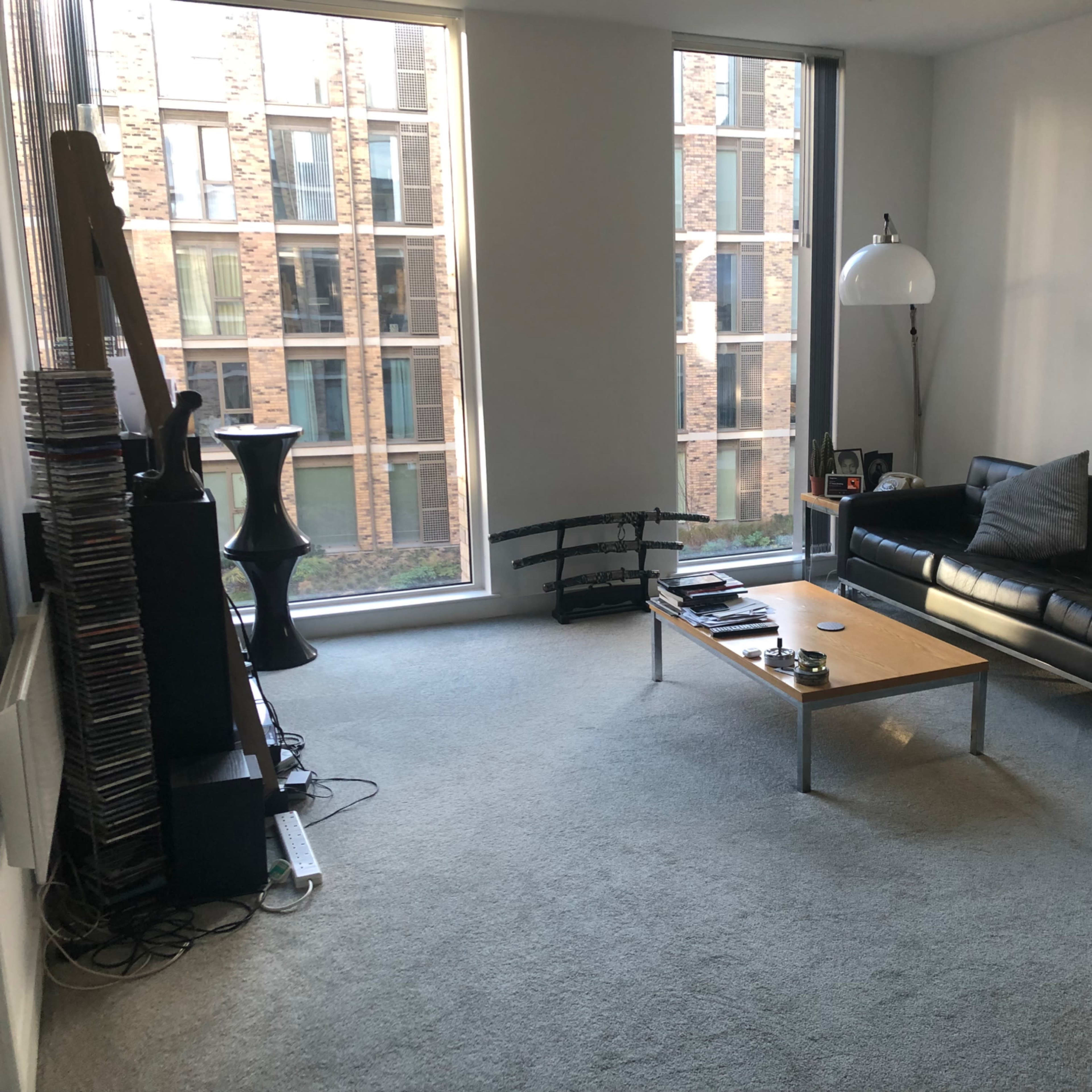A sparsely decorated living room with a black leather sofa, a wooden coffee table, and two large windows revealing a view of a brick building outside.