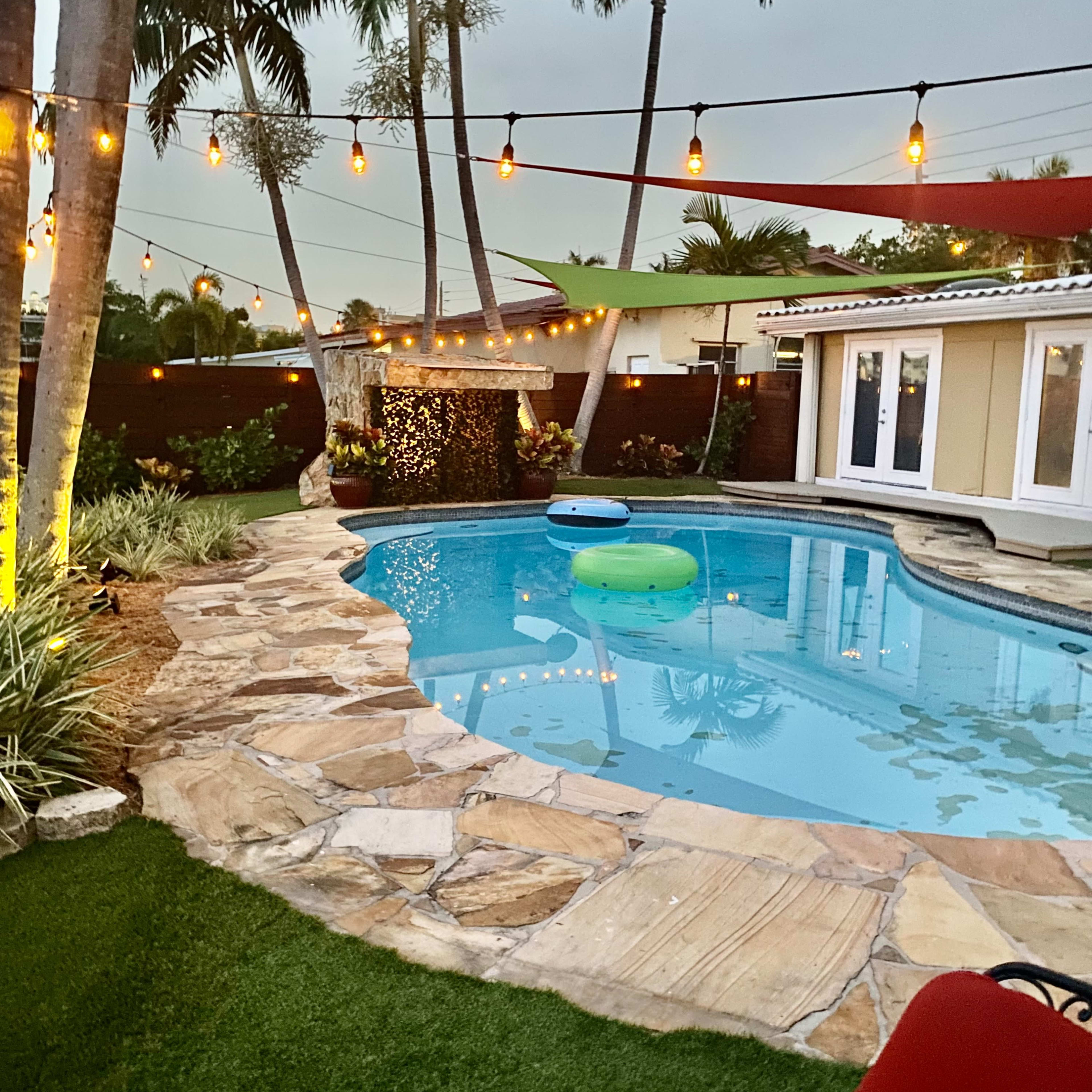 A pool area features a stone patio, green inflatable float, and palm trees adorned with string lights.