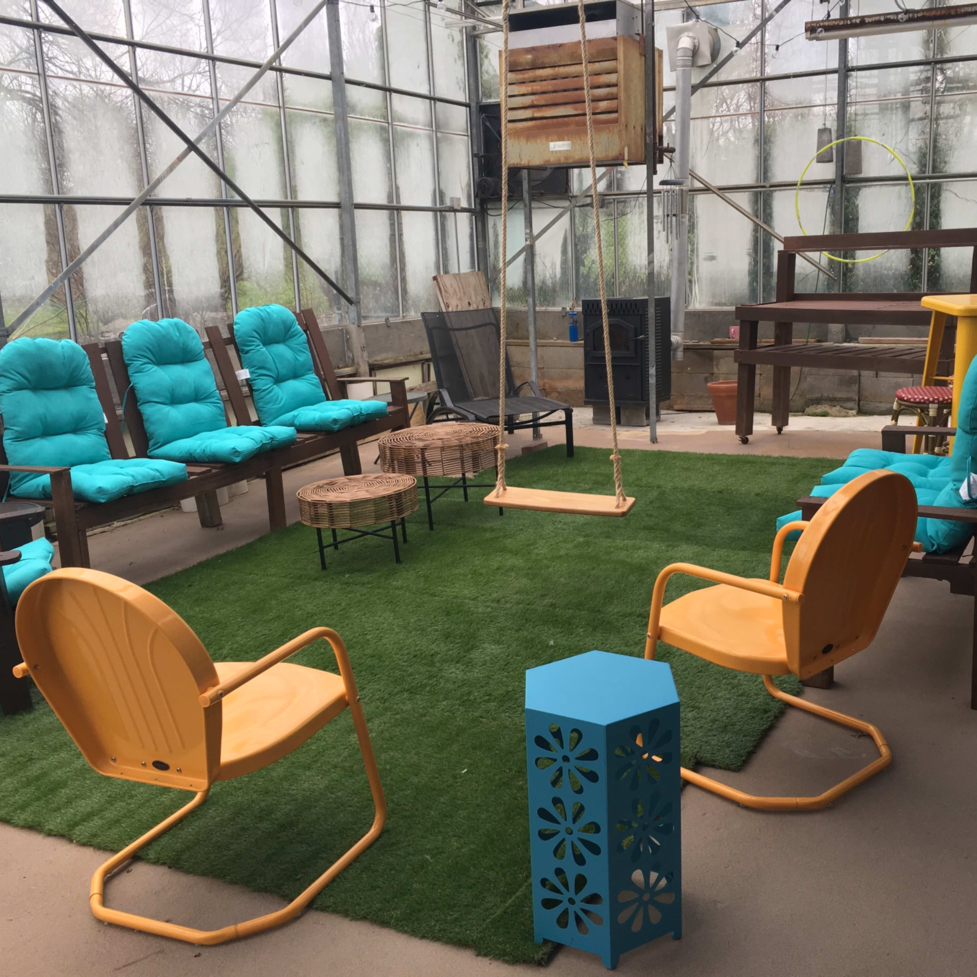 A greenhouse area furnished with bright chairs and a swing, arranged around a grassy carpet.