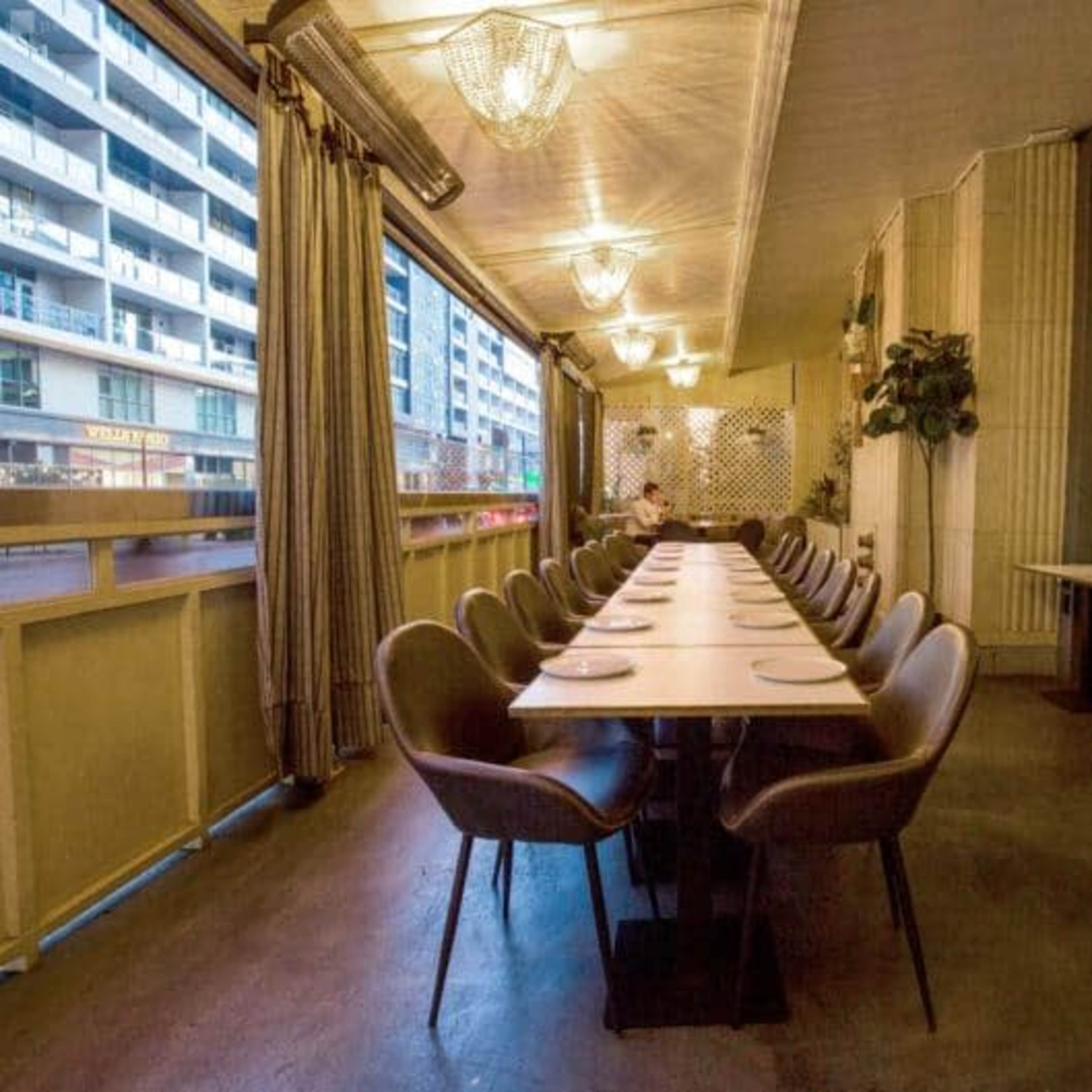 A long, empty dining table with chairs is set in a well-lit restaurant space that features large windows and modern decor.
