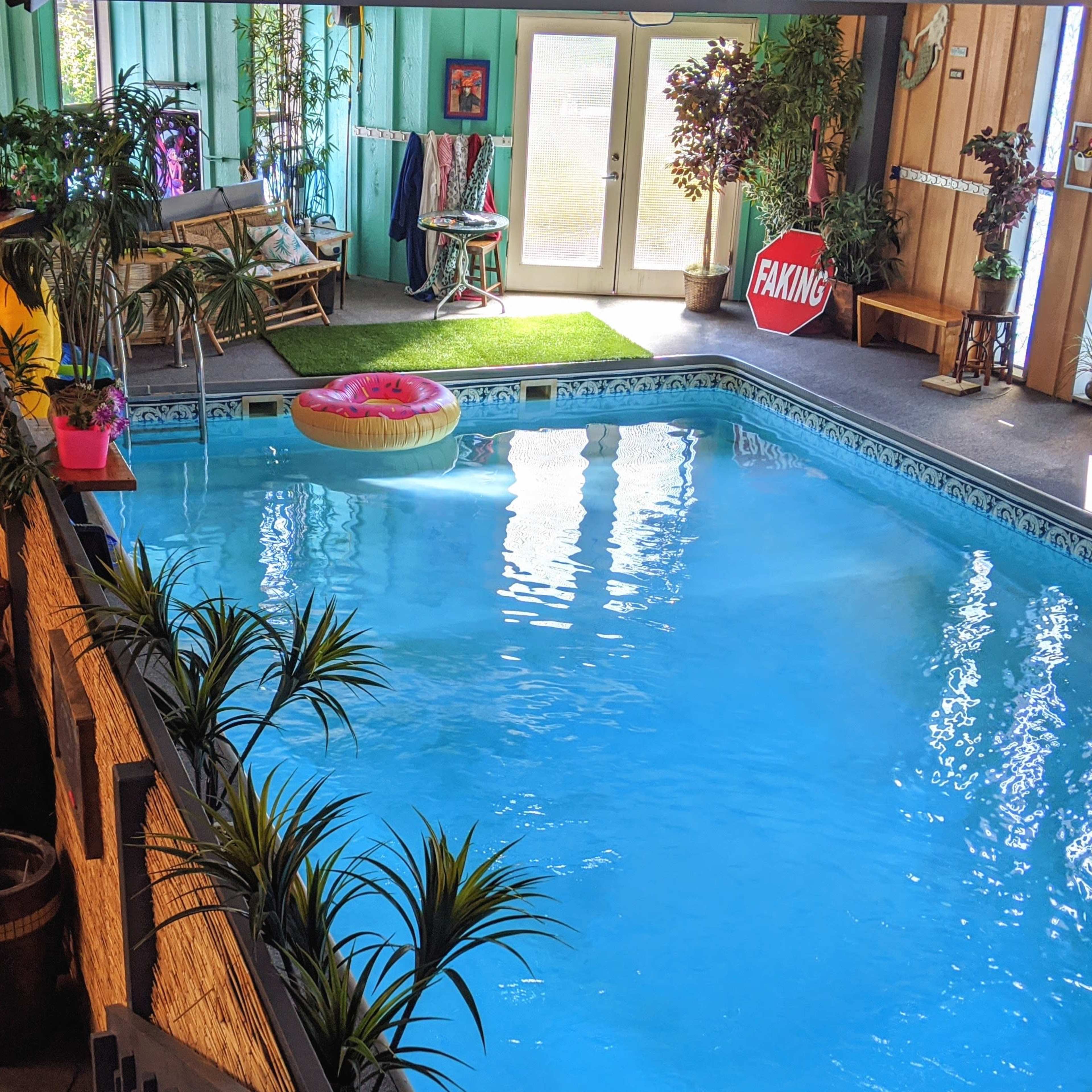 An indoor swimming pool surrounded by plants, a pool float, and lounge chairs, with large windows allowing natural light to enter.