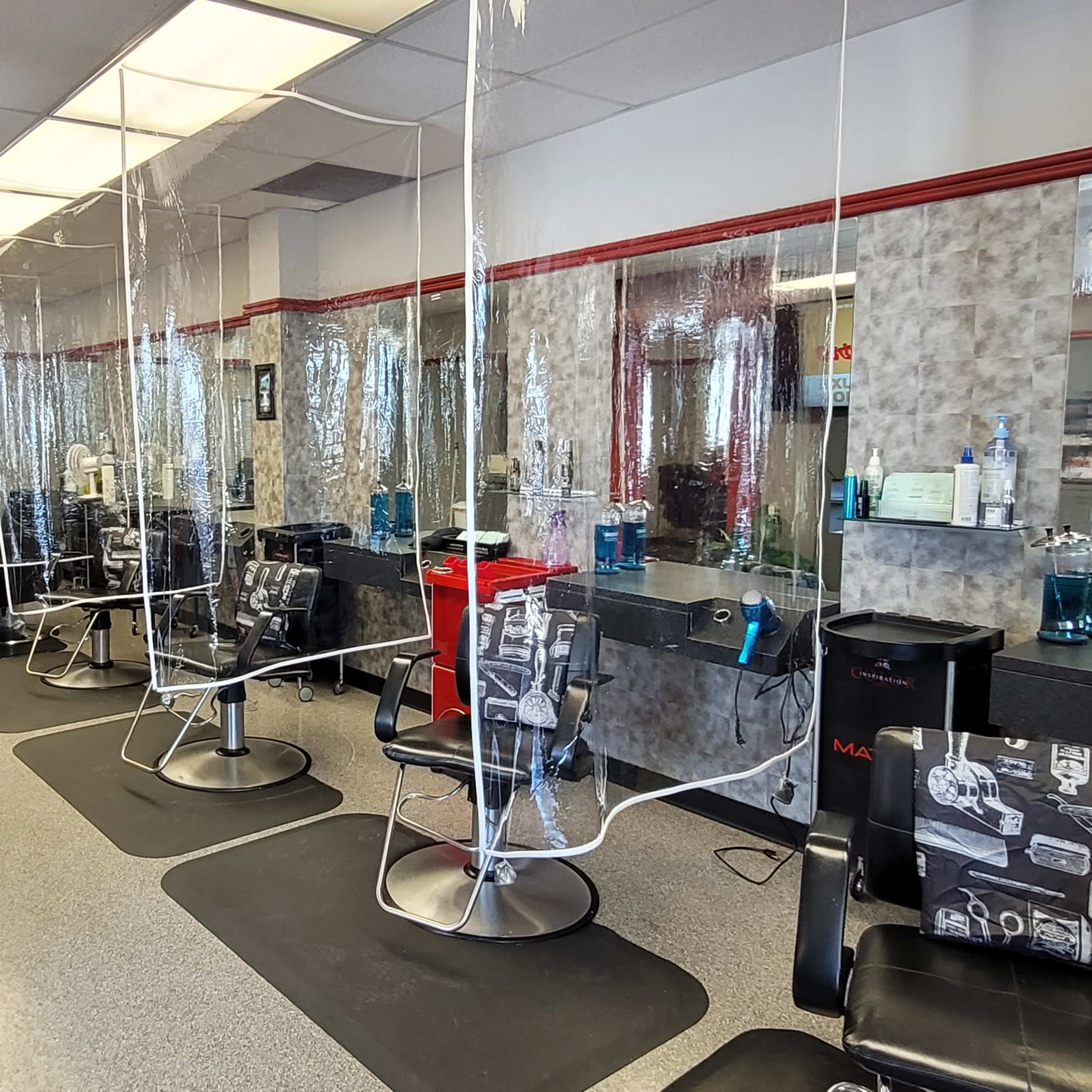 A hair salon interior features several styling stations with clear plastic dividers and black barber chairs arranged along a mirrored wall.
