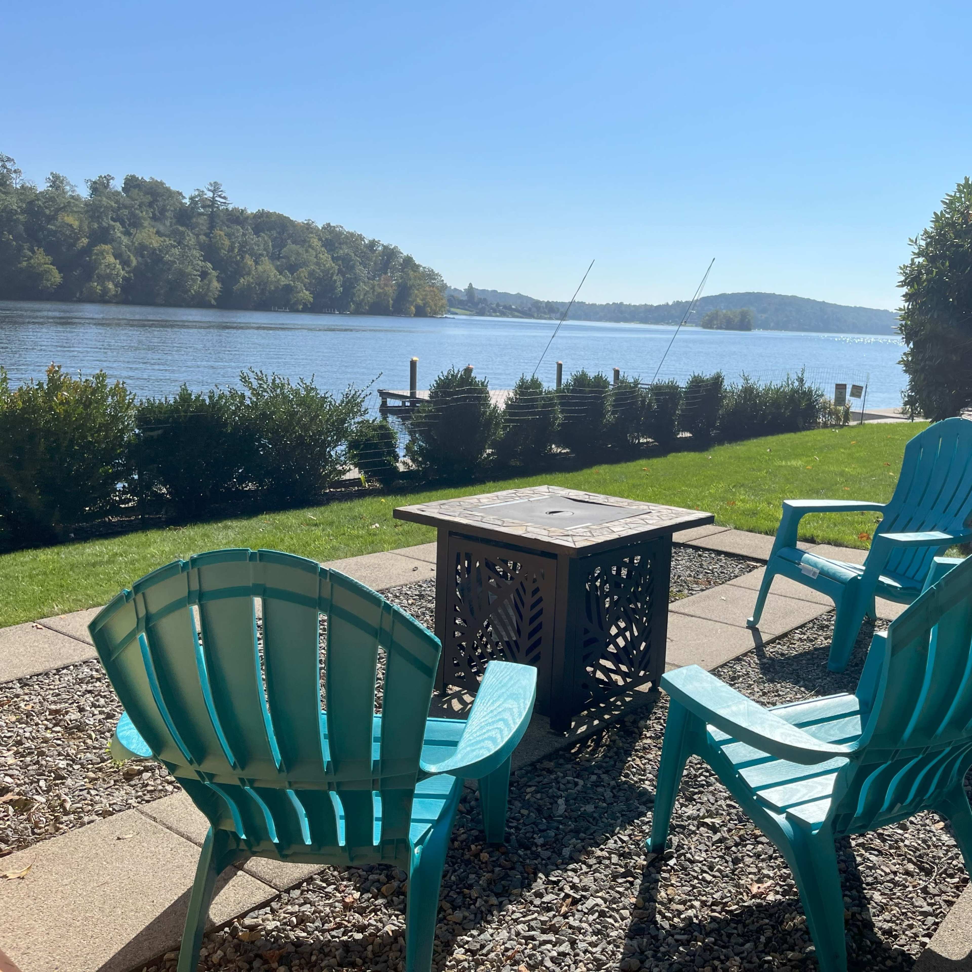A view of four teal Adirondack chairs arranged around a fire pit, facing a tranquil lake surrounded by greenery.