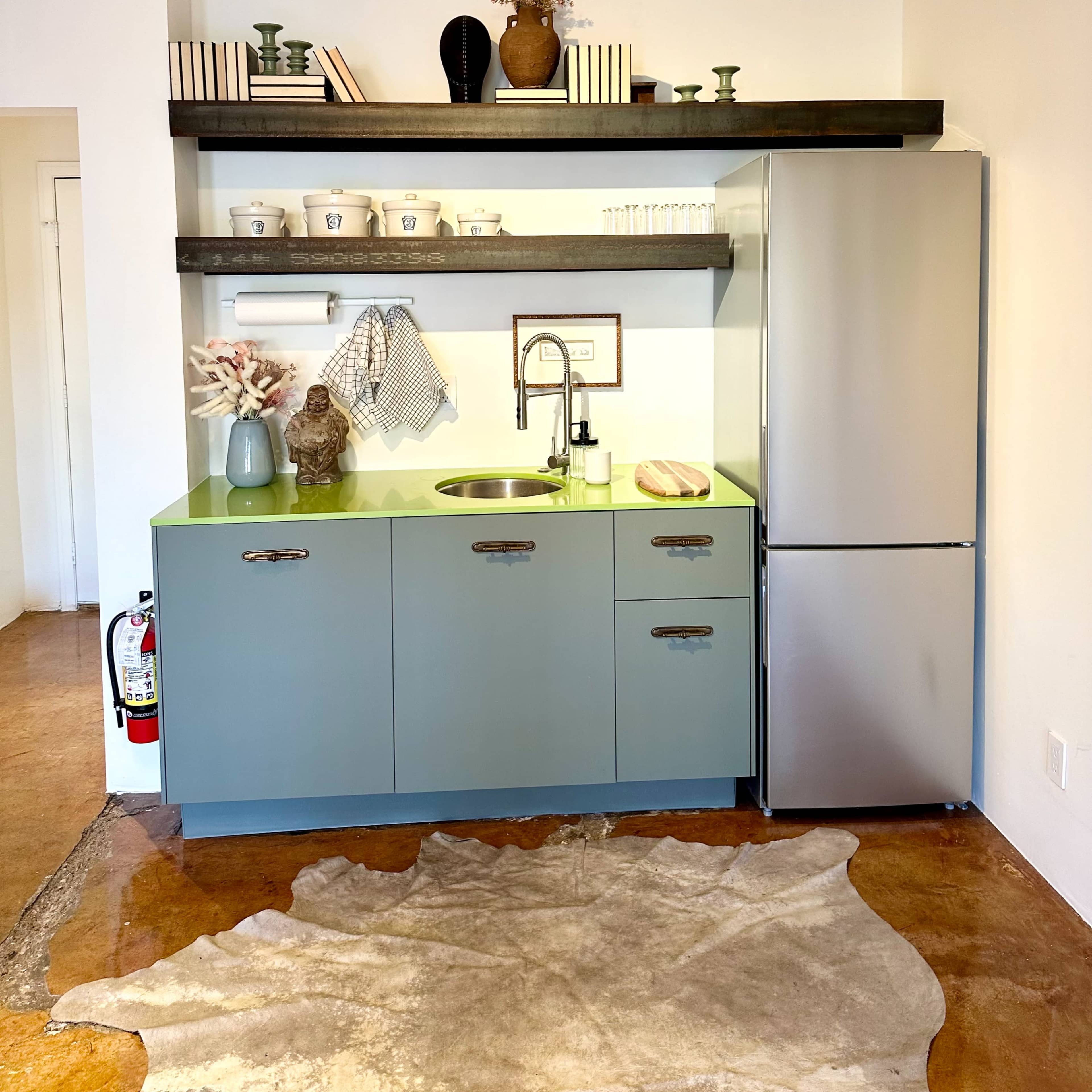 A minimalist kitchen features a green countertop, a stainless steel sink, a refrigerator, and an open shelf displaying decorative items.