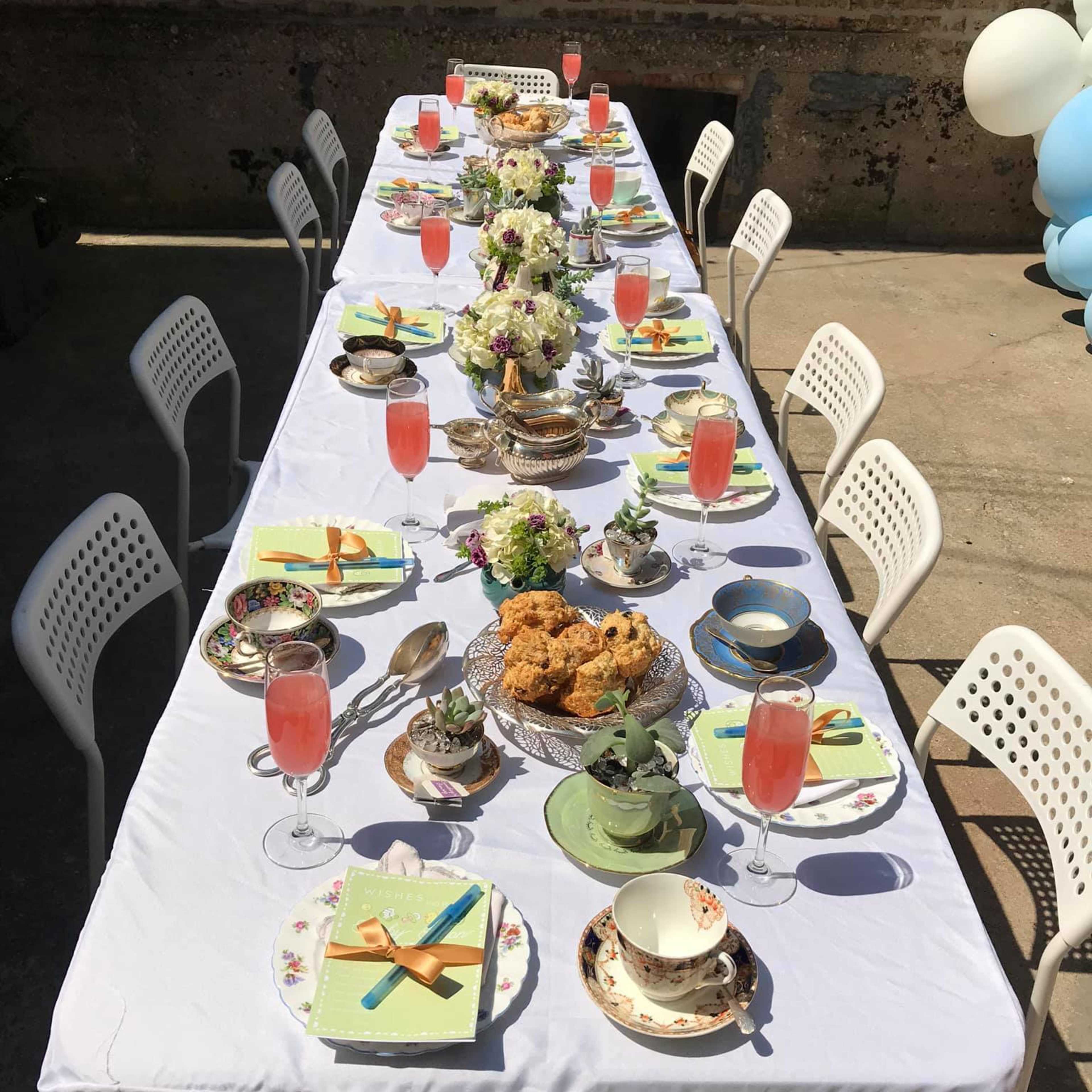 A long table is set outdoors with colorful tableware, floral centerpieces, and drinks in glasses, arranged for a gathering.