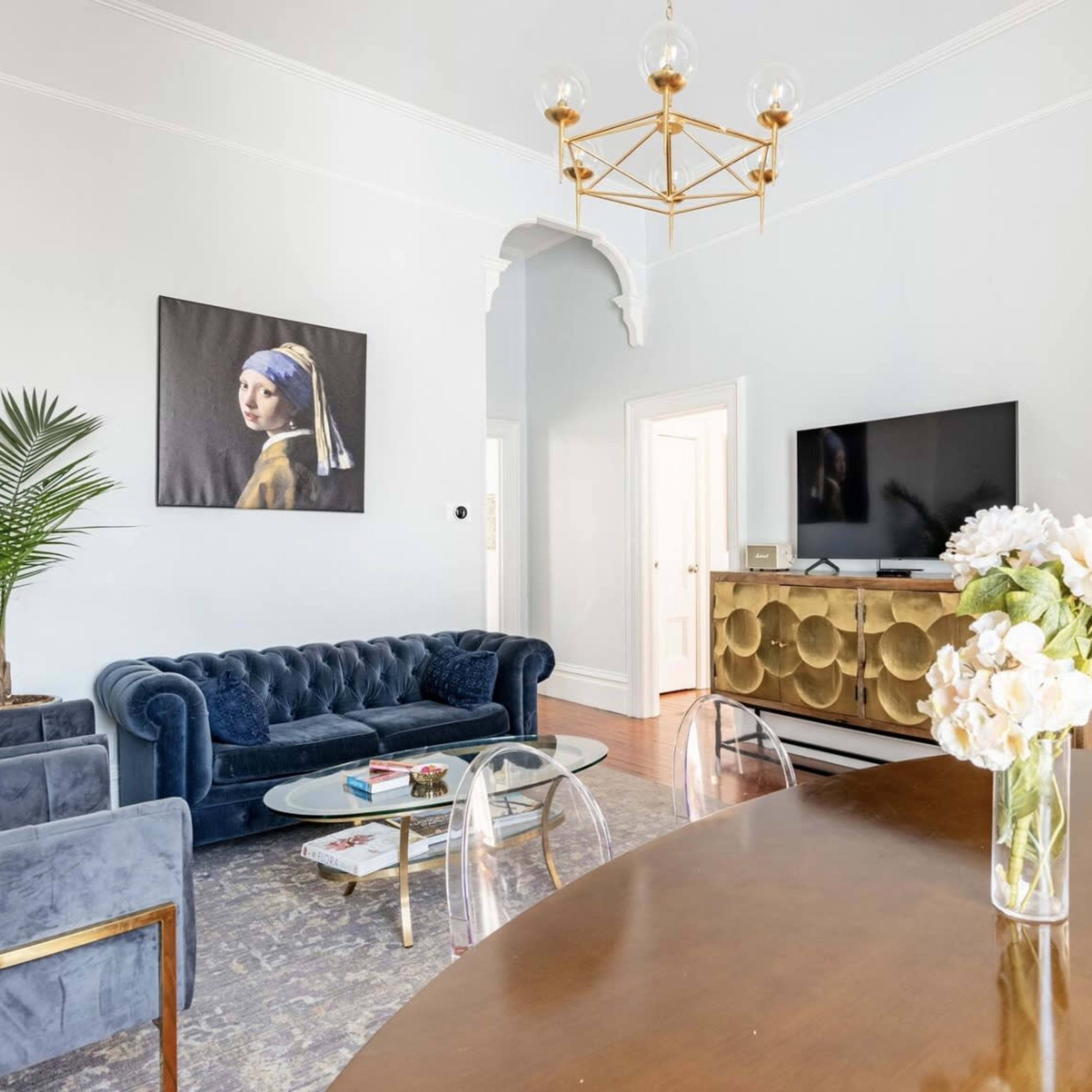 A bright living room featuring a blue velvet sofa, a coffee table, a flat-screen TV on a gold console, and a dining table with a vase of white flowers.