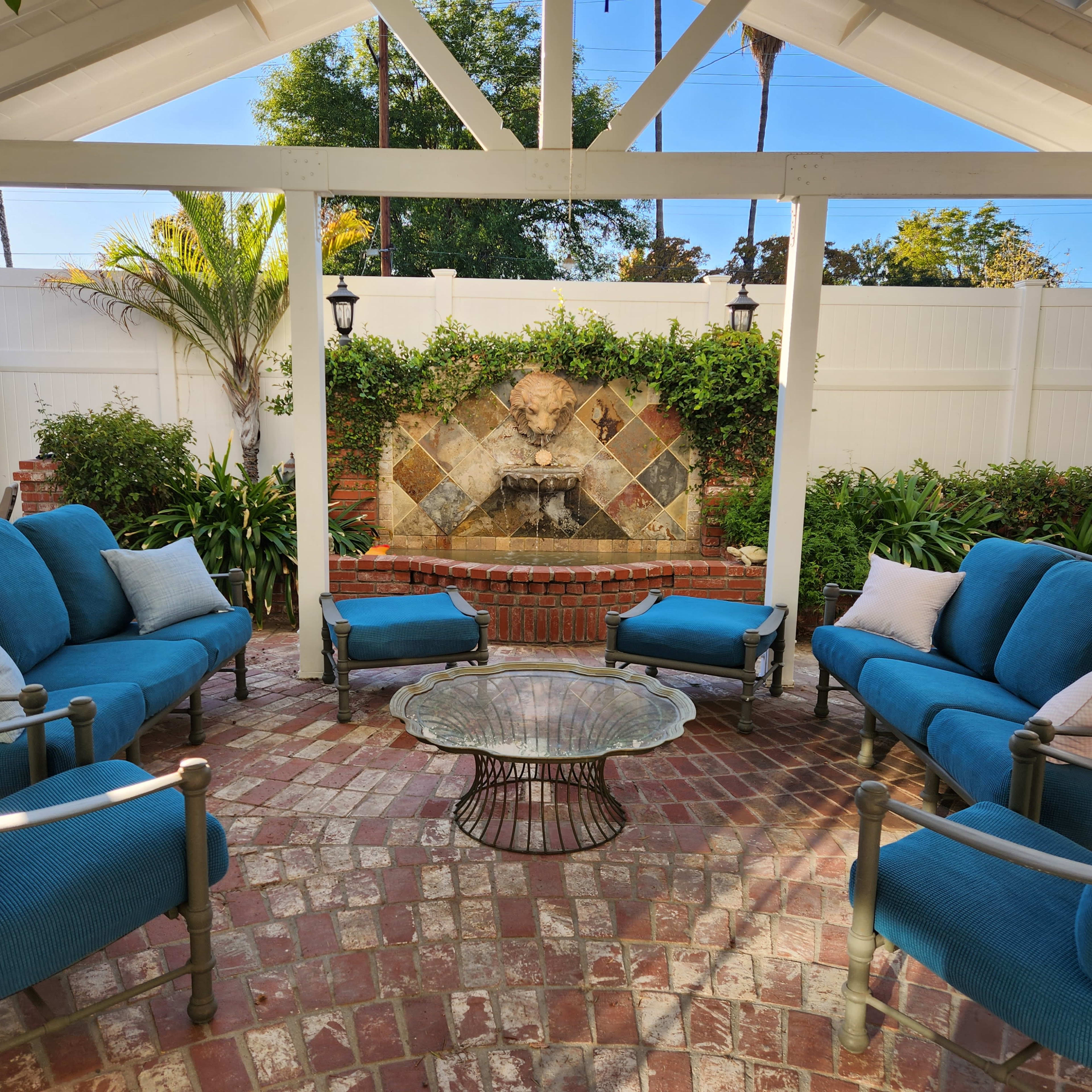 A shaded outdoor seating area with blue sofas, a glass table, and a wall fountain surrounded by greenery.