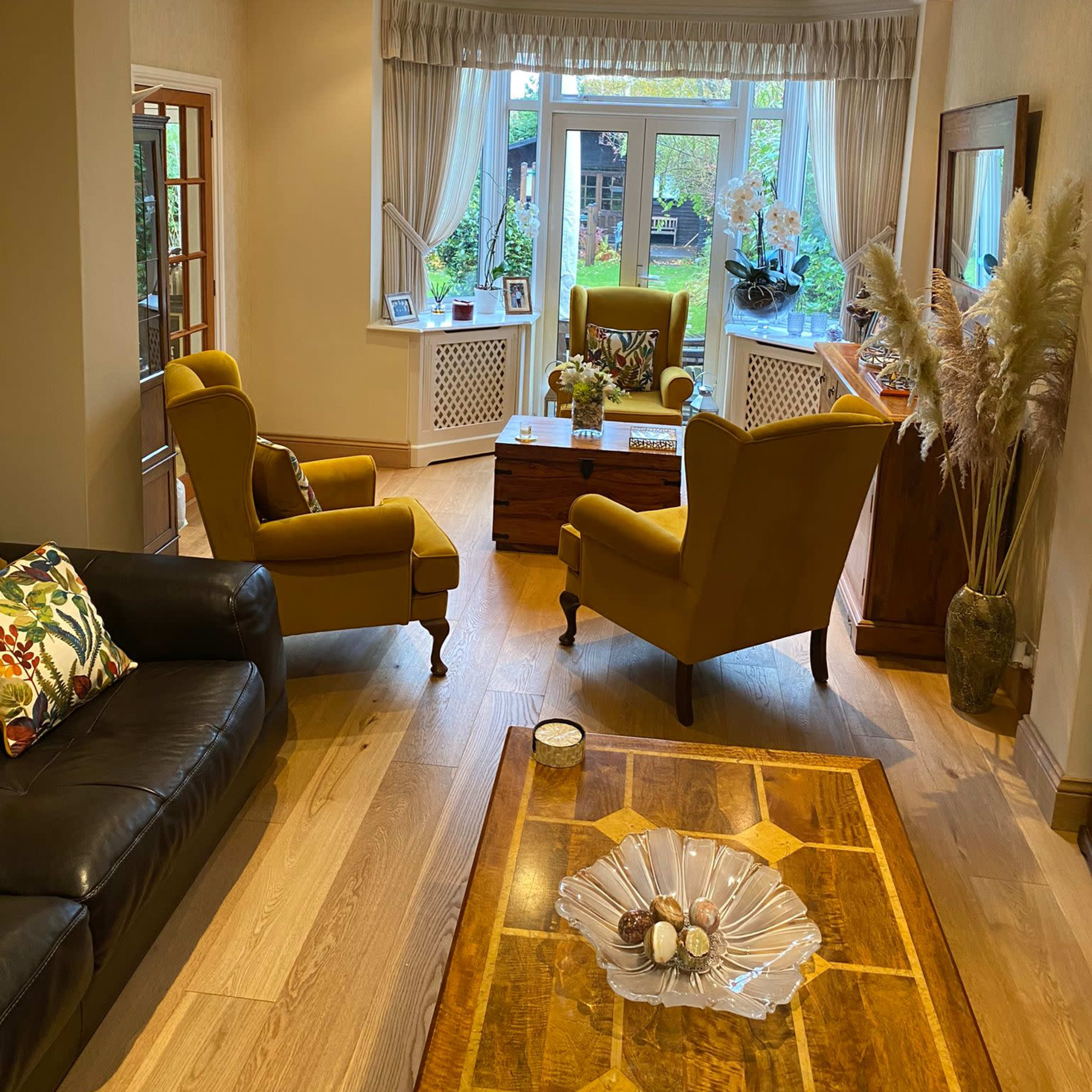 A living room with two yellow armchairs facing a wooden coffee table, a dark sofa, and large windows with a view of greenery outside.
