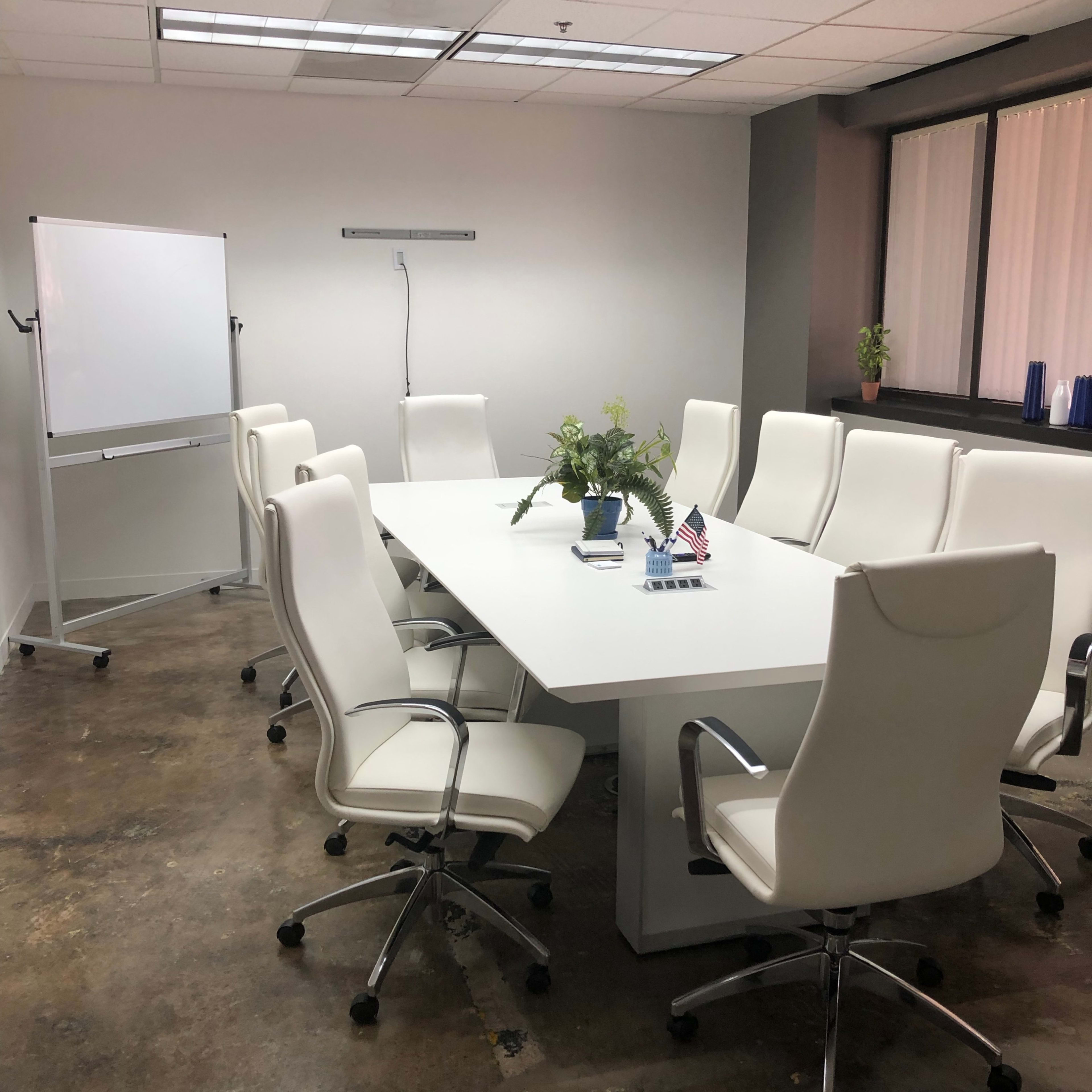 A conference room features a long white table surrounded by eight white chairs, with a flip chart and a small potted plant on the table.