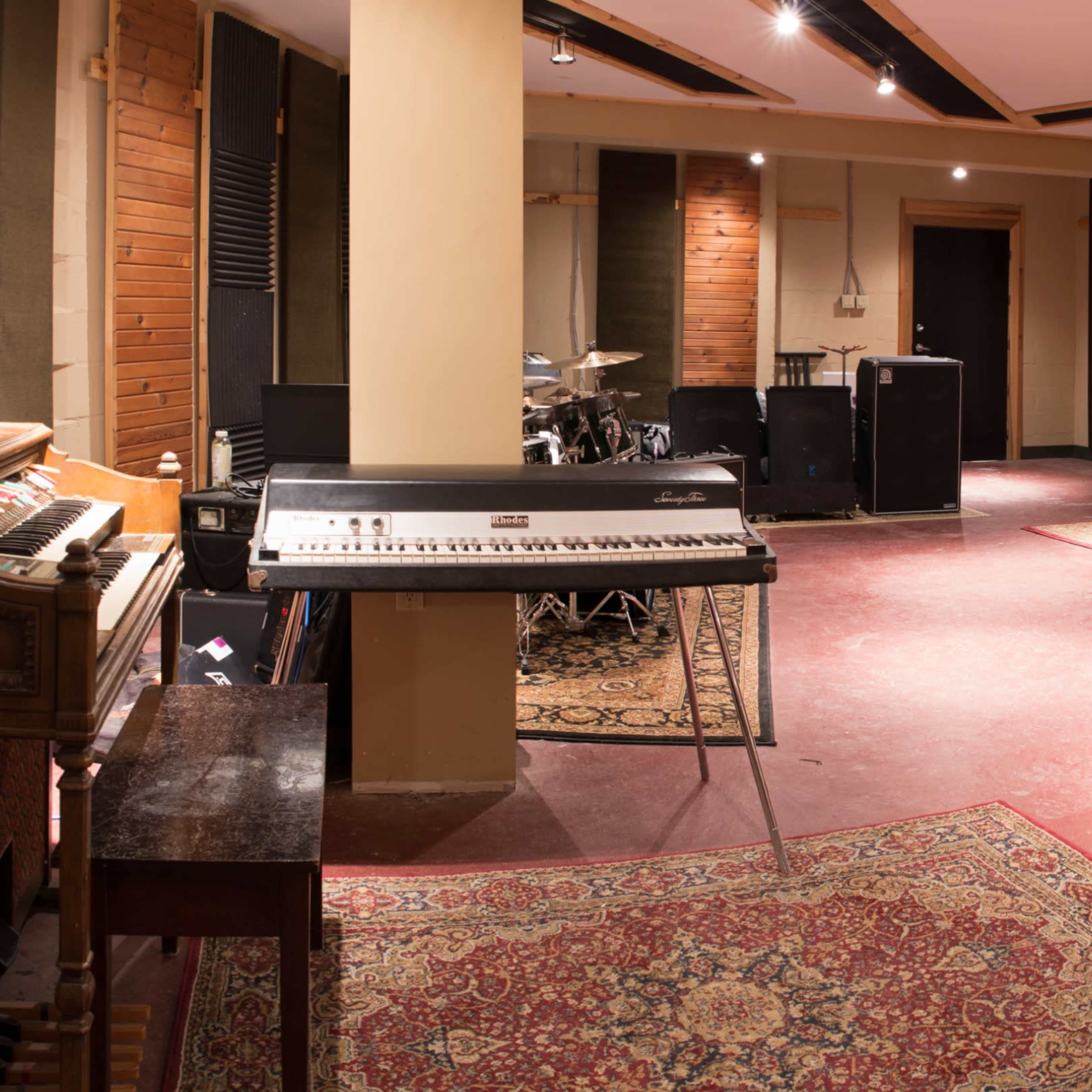 A spacious music studio featuring various instruments, amplifiers, and sound equipment arranged around a large, empty floor with decorative rugs.