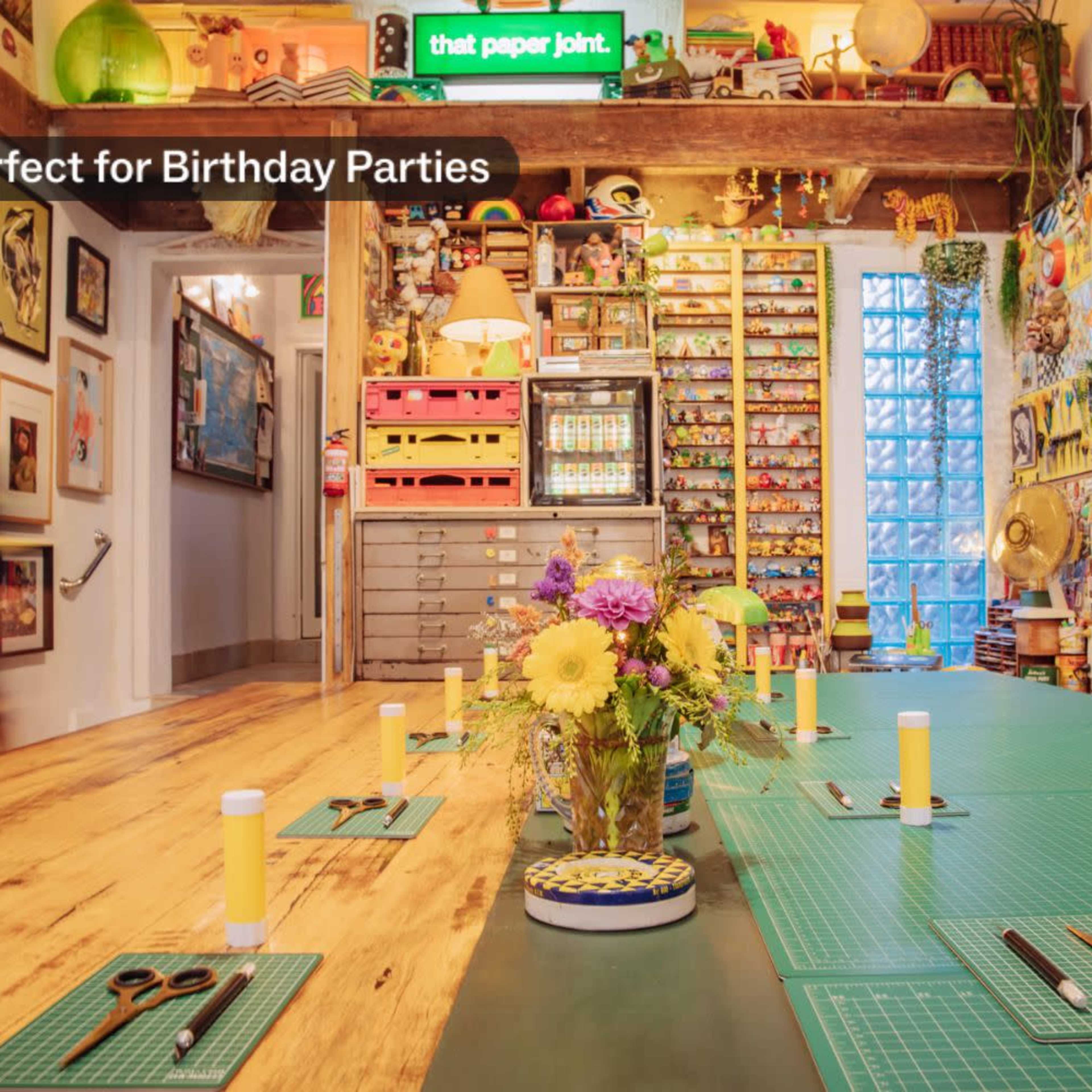 A bright, colorful crafting space is set up with a central flower arrangement and crafting supplies organized on tables, surrounded by walls adorned with various artworks.