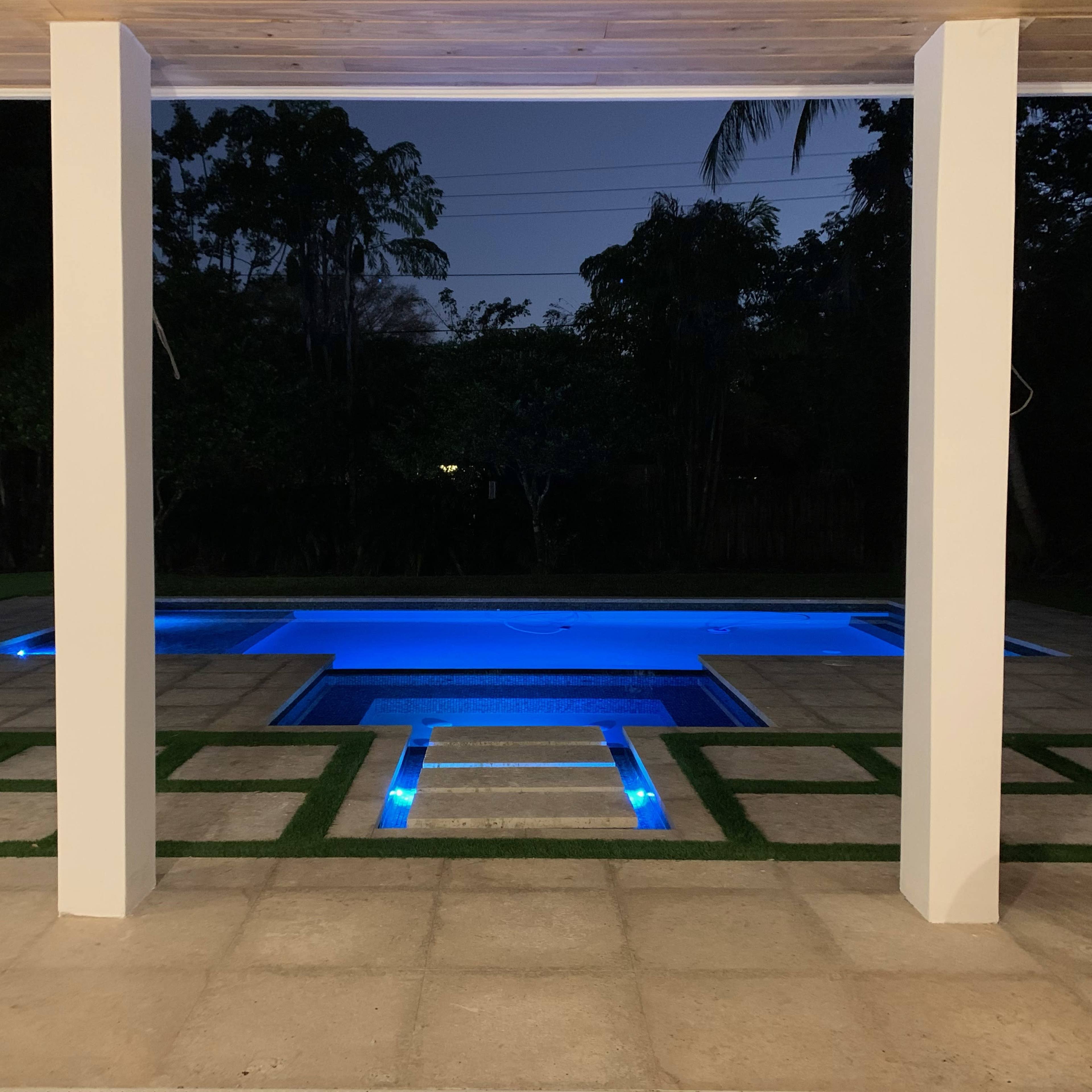 A well-lit swimming pool with blue illumination sits in a landscaped backyard at night, framed by white columns.