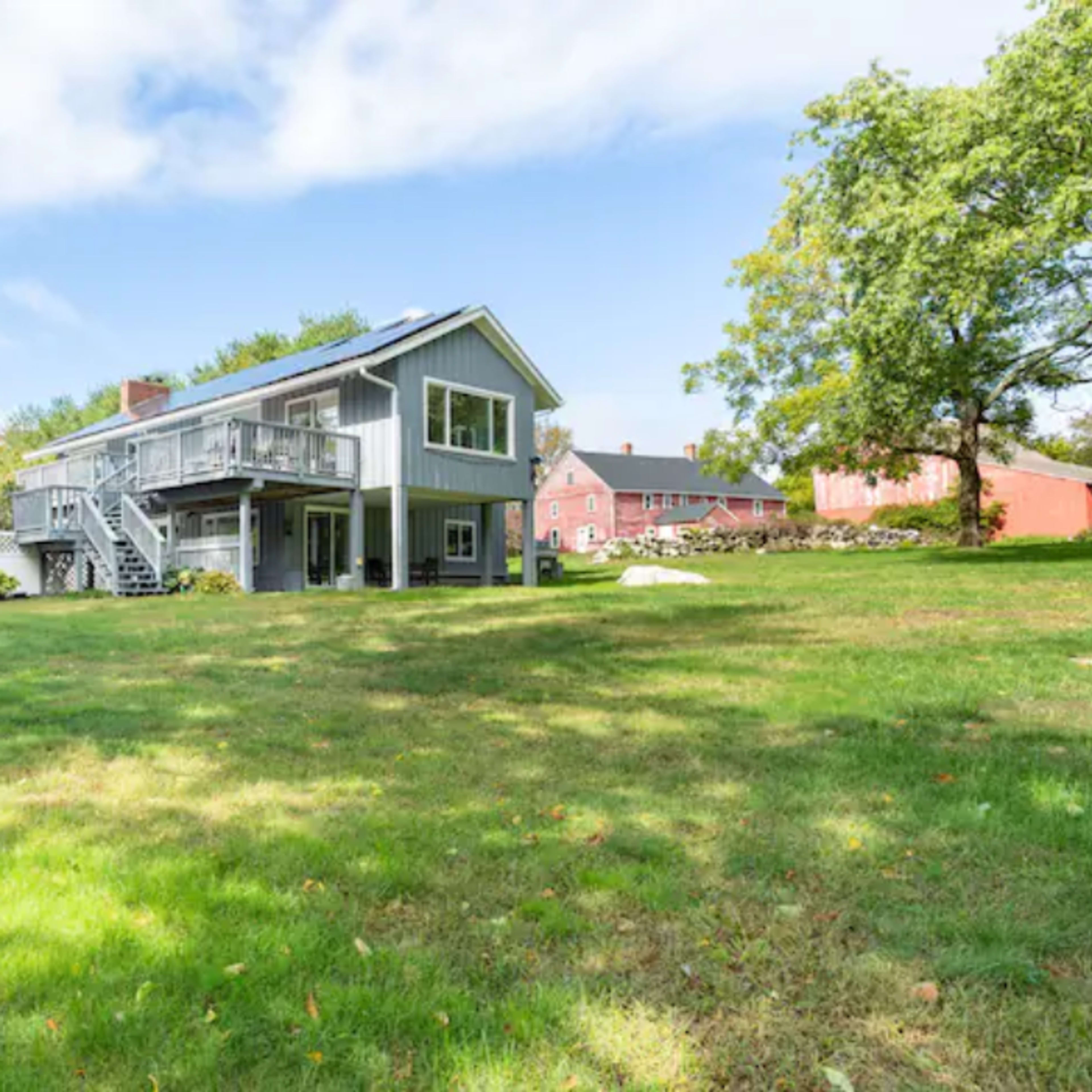 A two-story gray house sits on a grassy lawn, surrounded by trees and other buildings.