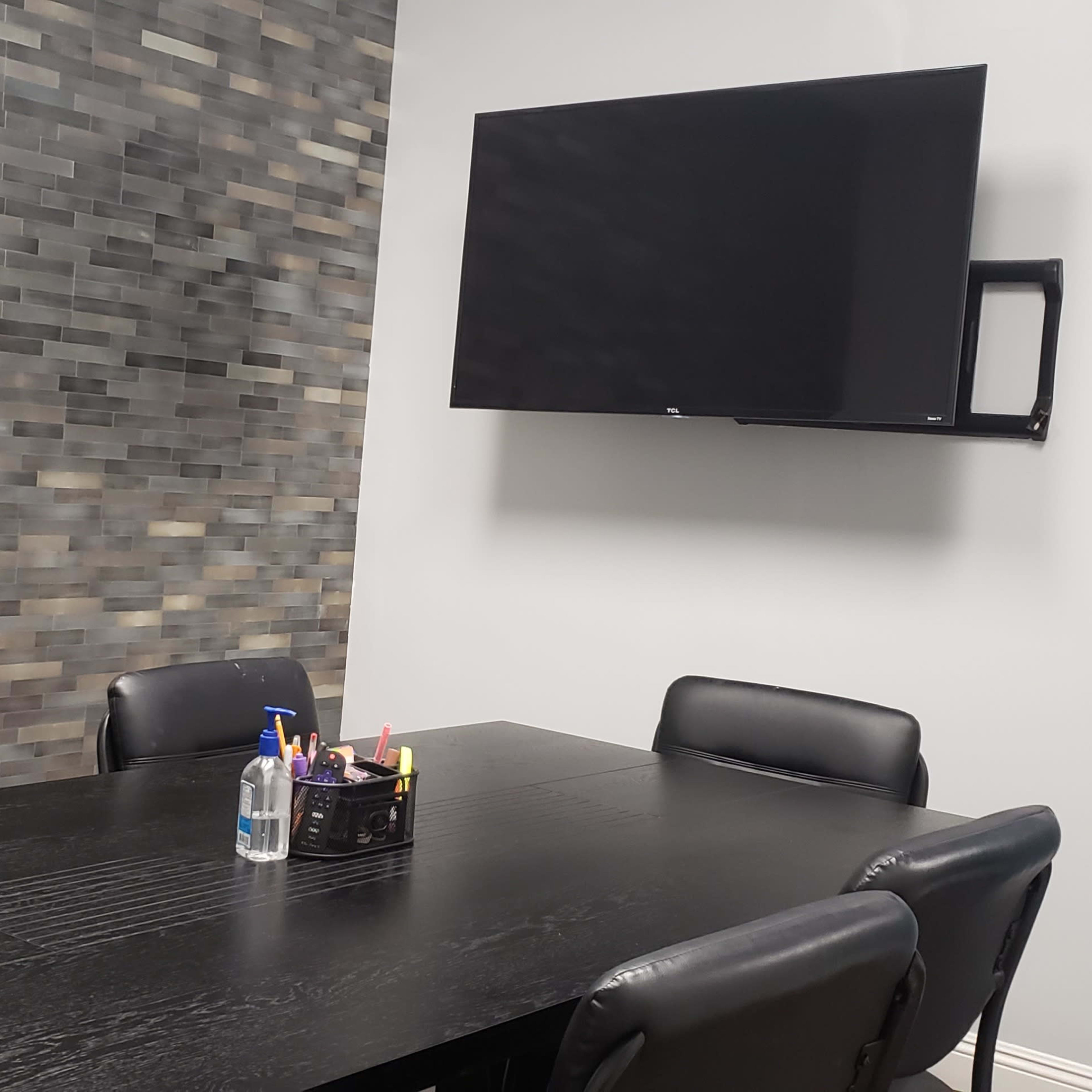A conference room features a black table surrounded by chairs, a wall-mounted television, and a stone-tiled accent wall.