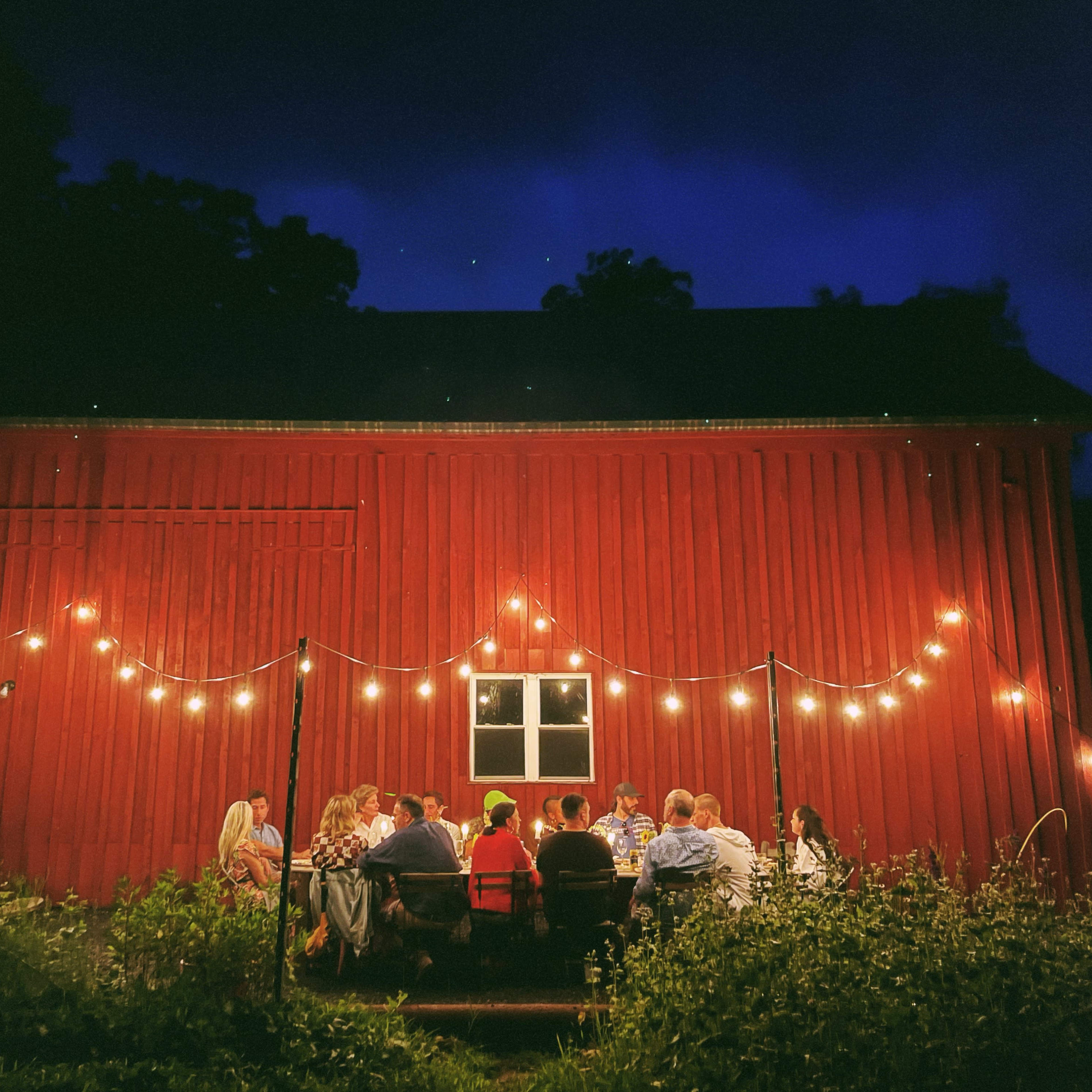 A group of people gathers around a table outside a red barn, illuminated by string lights against a darkening sky.