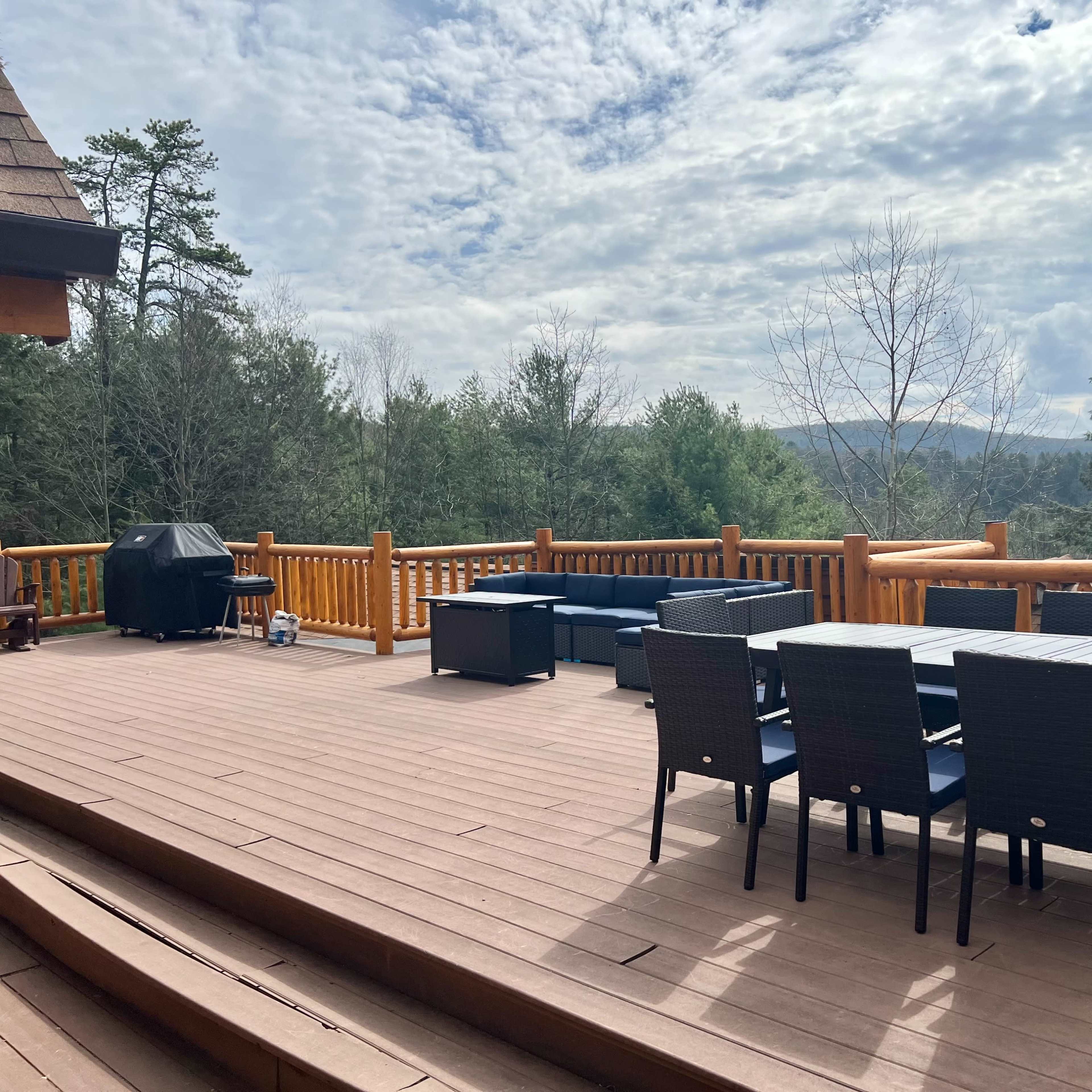 A wooden deck features a dining table with chairs, a grill, and seating areas, surrounded by trees and hills under a cloudy sky.