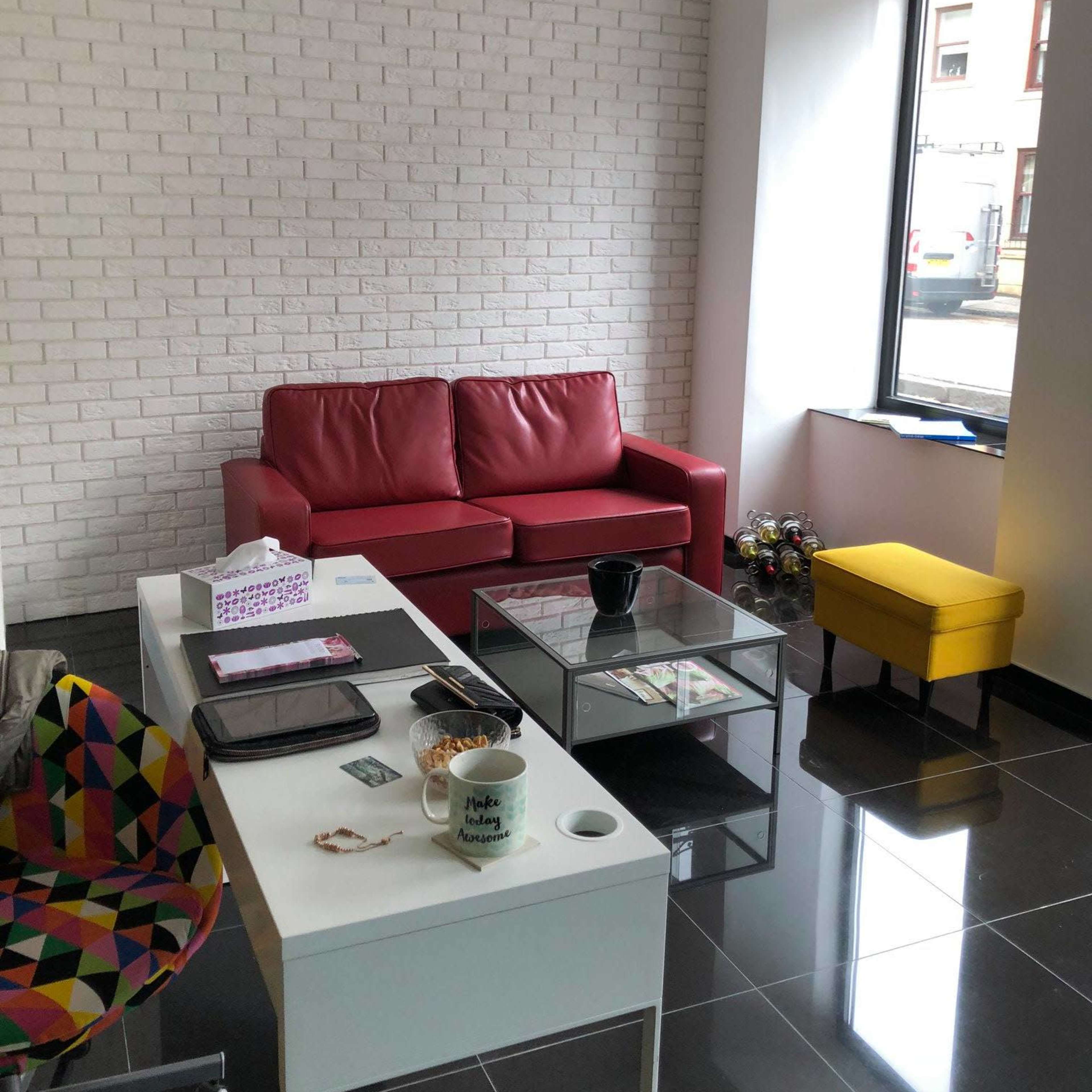 A modern living space with a red sofa, a yellow accent chair, and a glass coffee table, set against a white brick wall and black tiled floor.