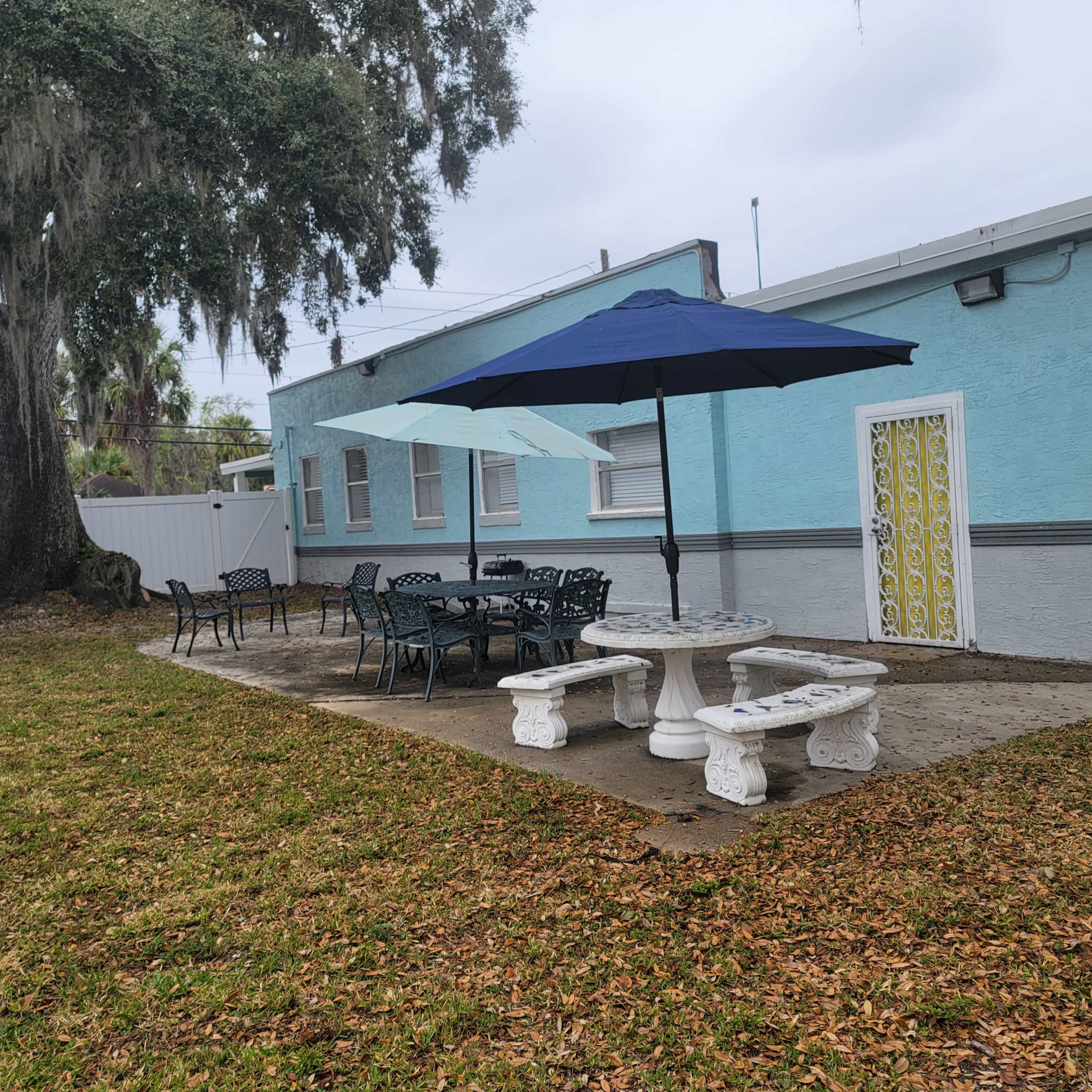 An outdoor seating area with a stone table and chairs under blue umbrellas beside a light blue building.