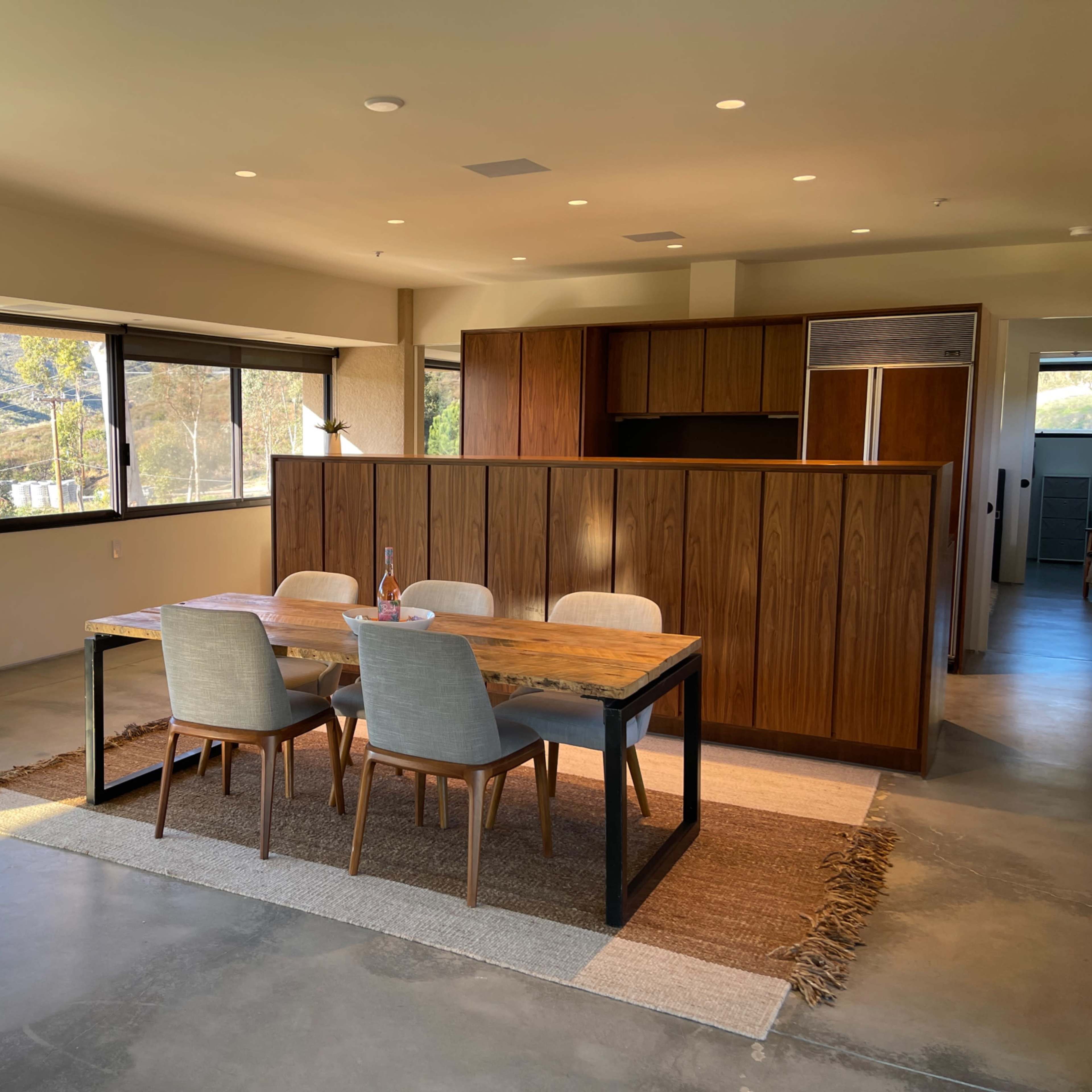 A modern dining area with a wooden table and four chairs, adjacent to an open kitchen, featuring large windows that offer views of a mountainous landscape.