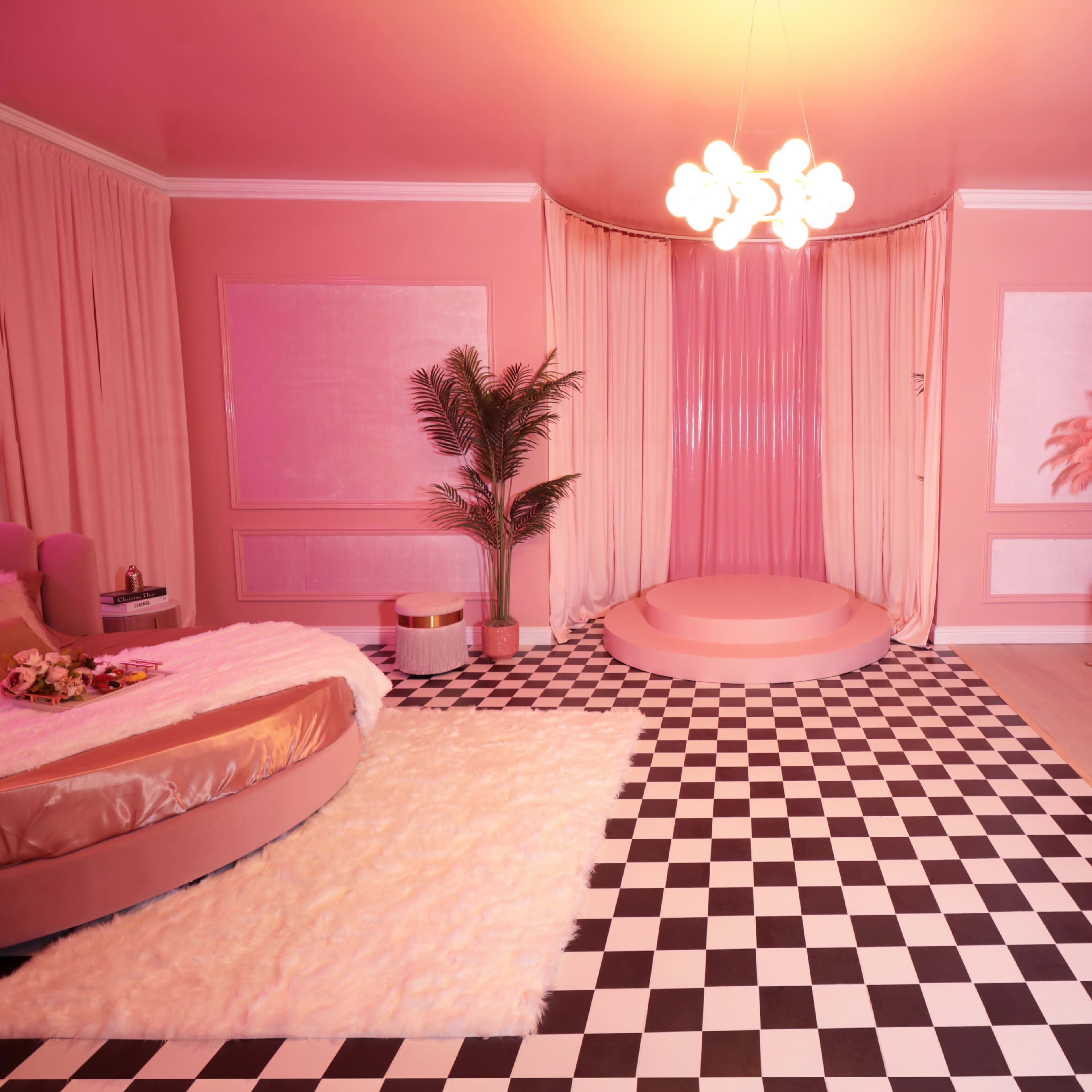 A pink-themed room features a circular stage, a checkered floor, and a white desk with shelving filled with books and decorative items.