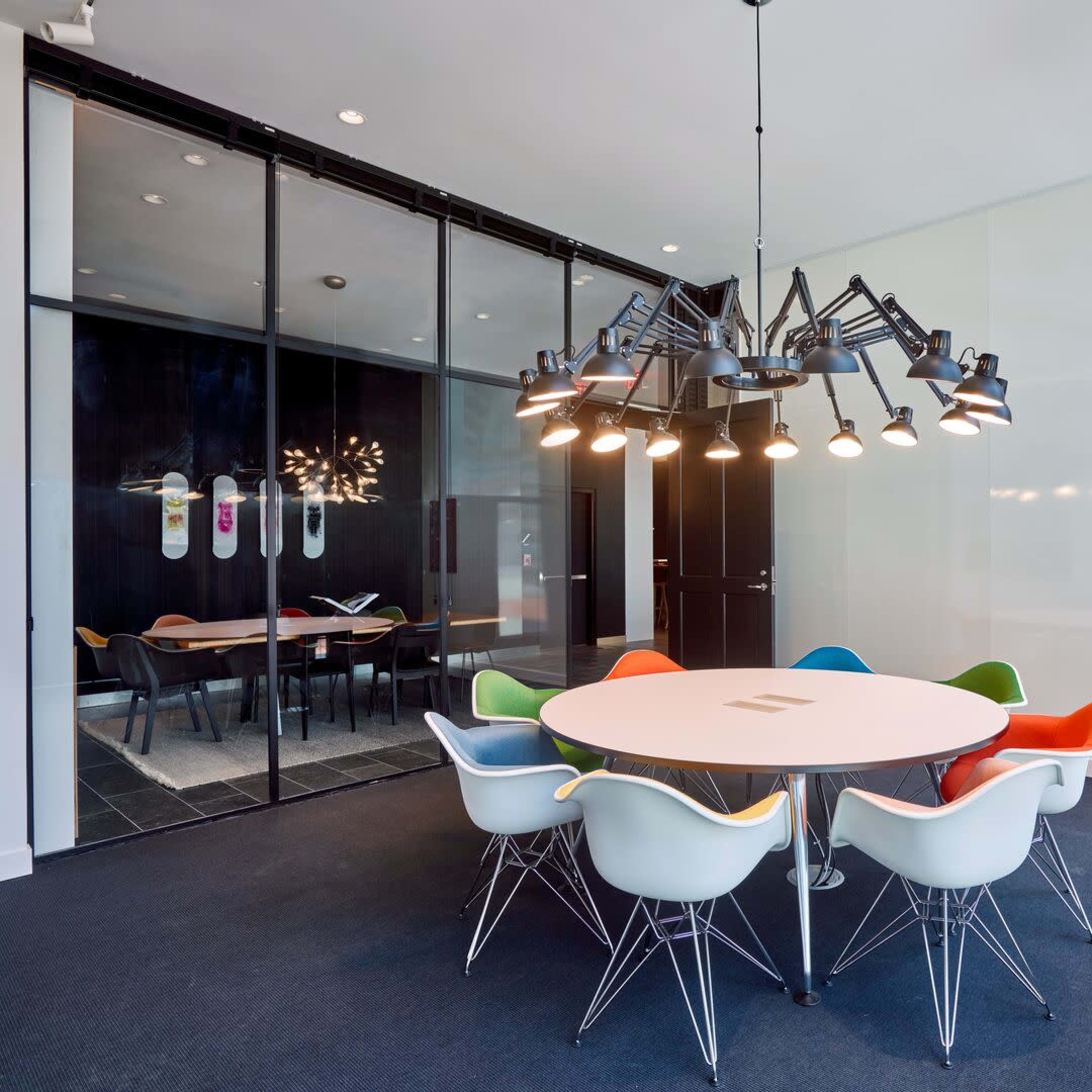 A modern conference room features a round table surrounded by colorful chairs and a large chandelier, with a glass wall revealing another meeting area.