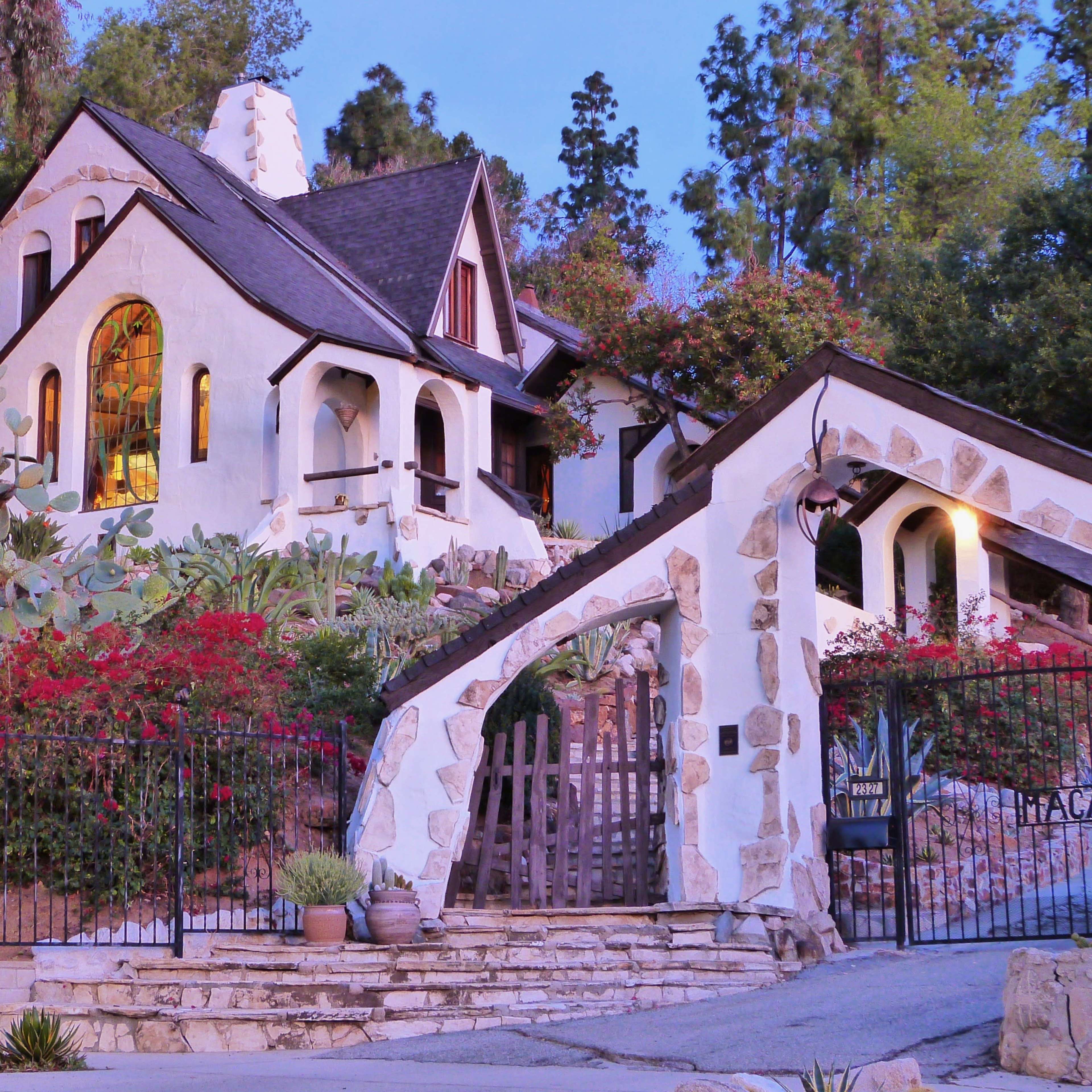 A white, textured house with a steep roof and arched windows is surrounded by lush greenery and colorful flowers.