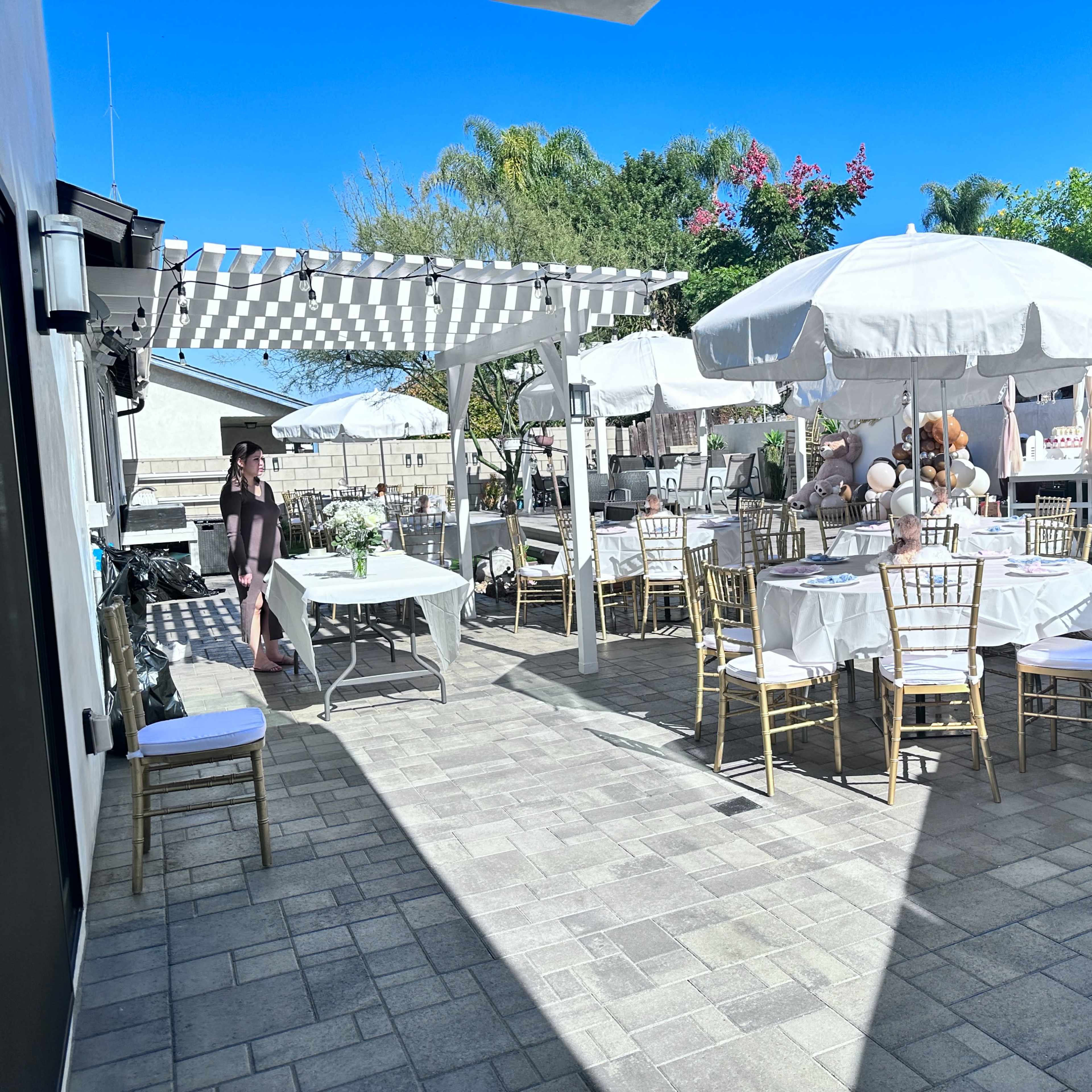 An outdoor patio decorated for an event, featuring tables with white tablecloths, golden chairs, umbrellas, and a pergola draped overhead.