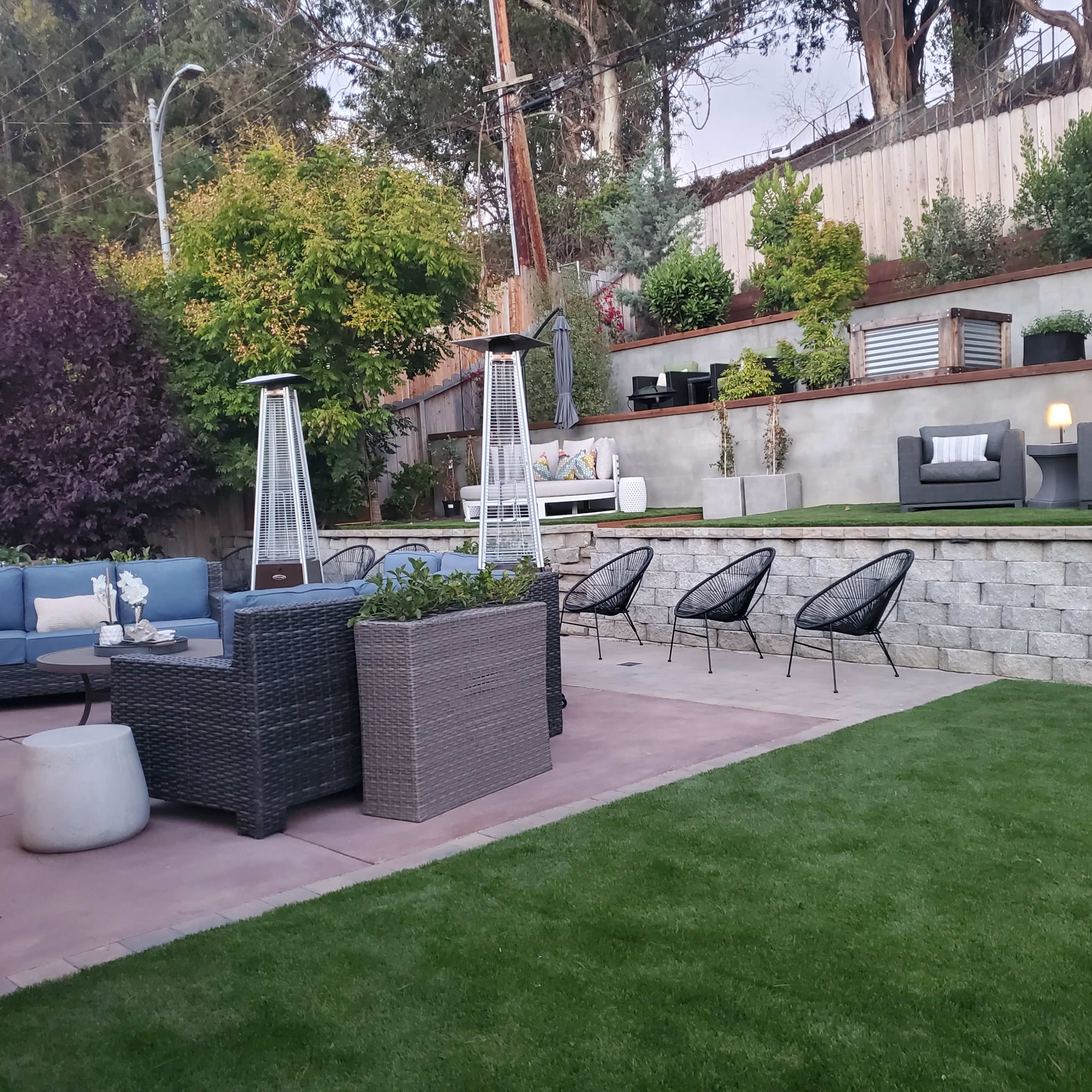 A landscaped backyard with seating areas, including a sofa set, lounge chairs, and a fire pit, bordered by greenery and stone walls.