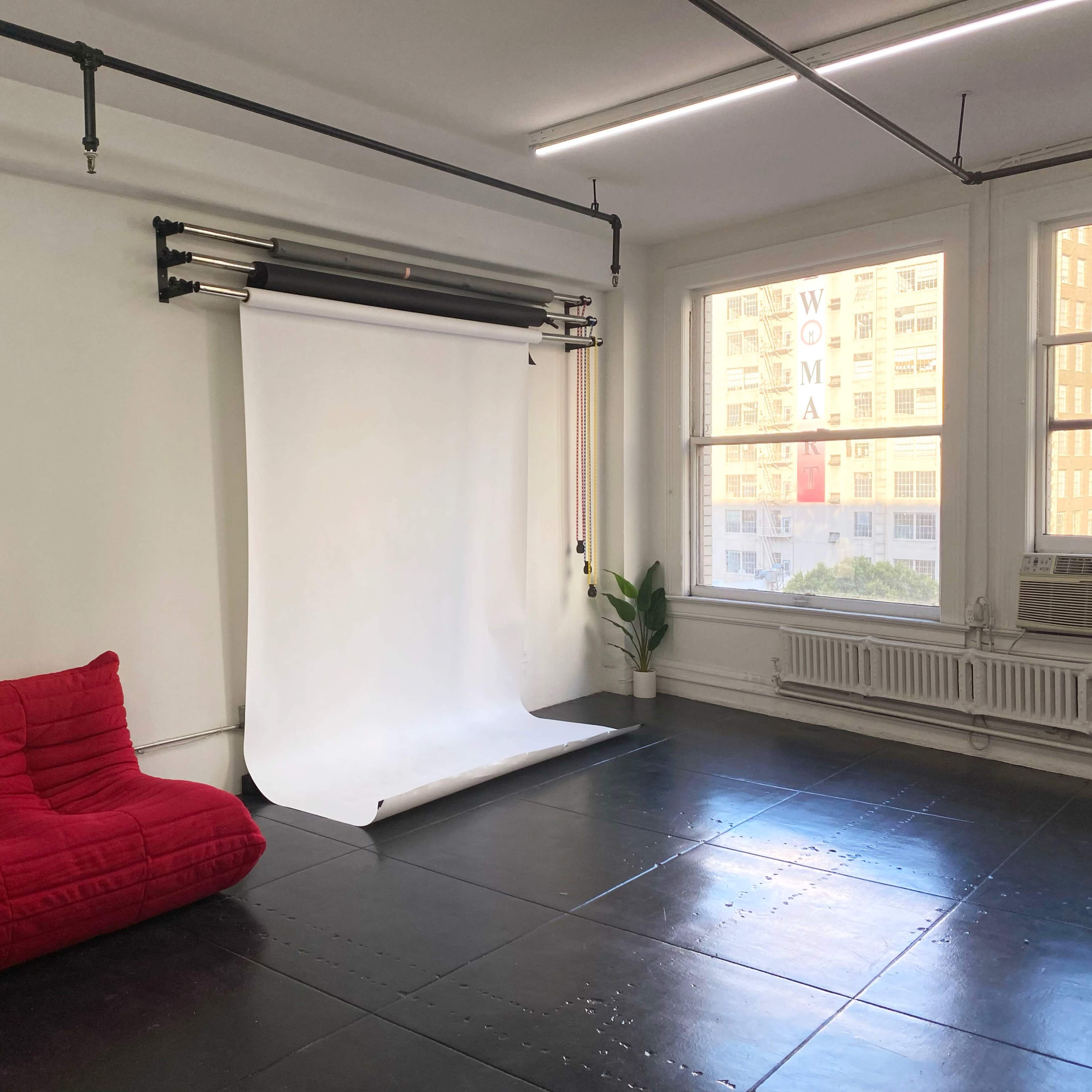A minimalist studio space with a red sofa, a white backdrop setup, and large windows letting in natural light.