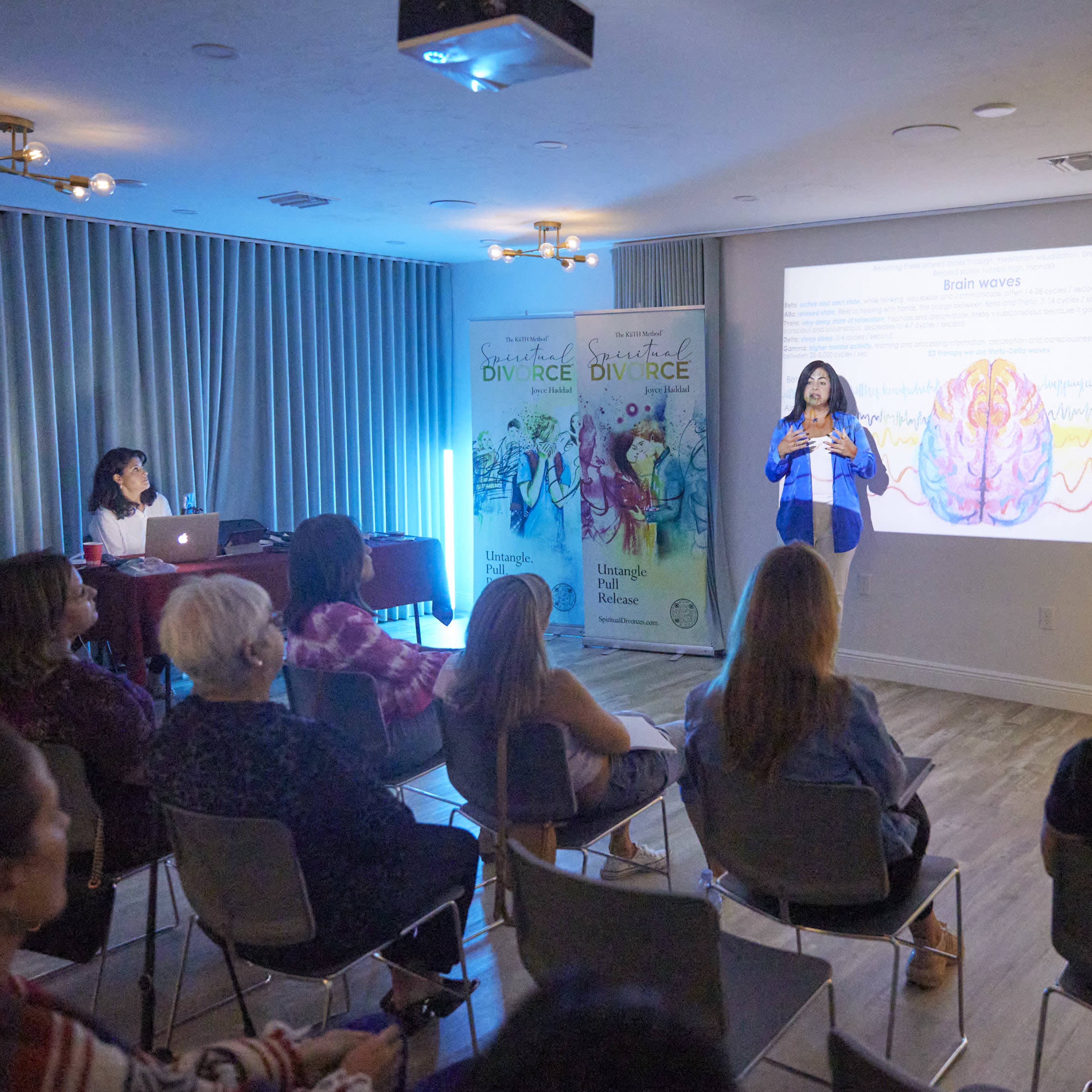 A speaker presents information on brain waves to an audience seated in a modern conference room with banners in the background.