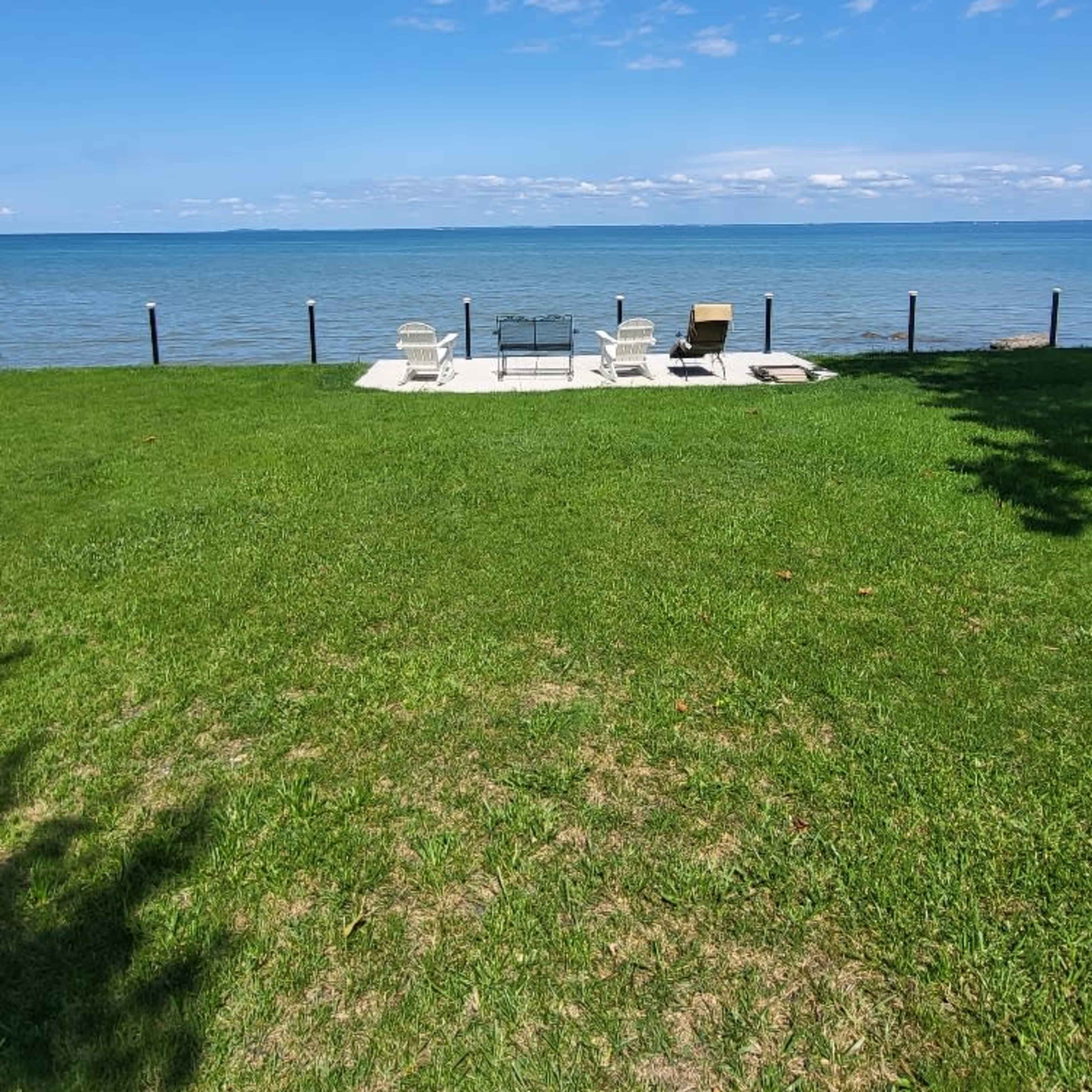 A patio with deck chairs and a table is situated on a grassy area facing a calm lake under a blue sky.