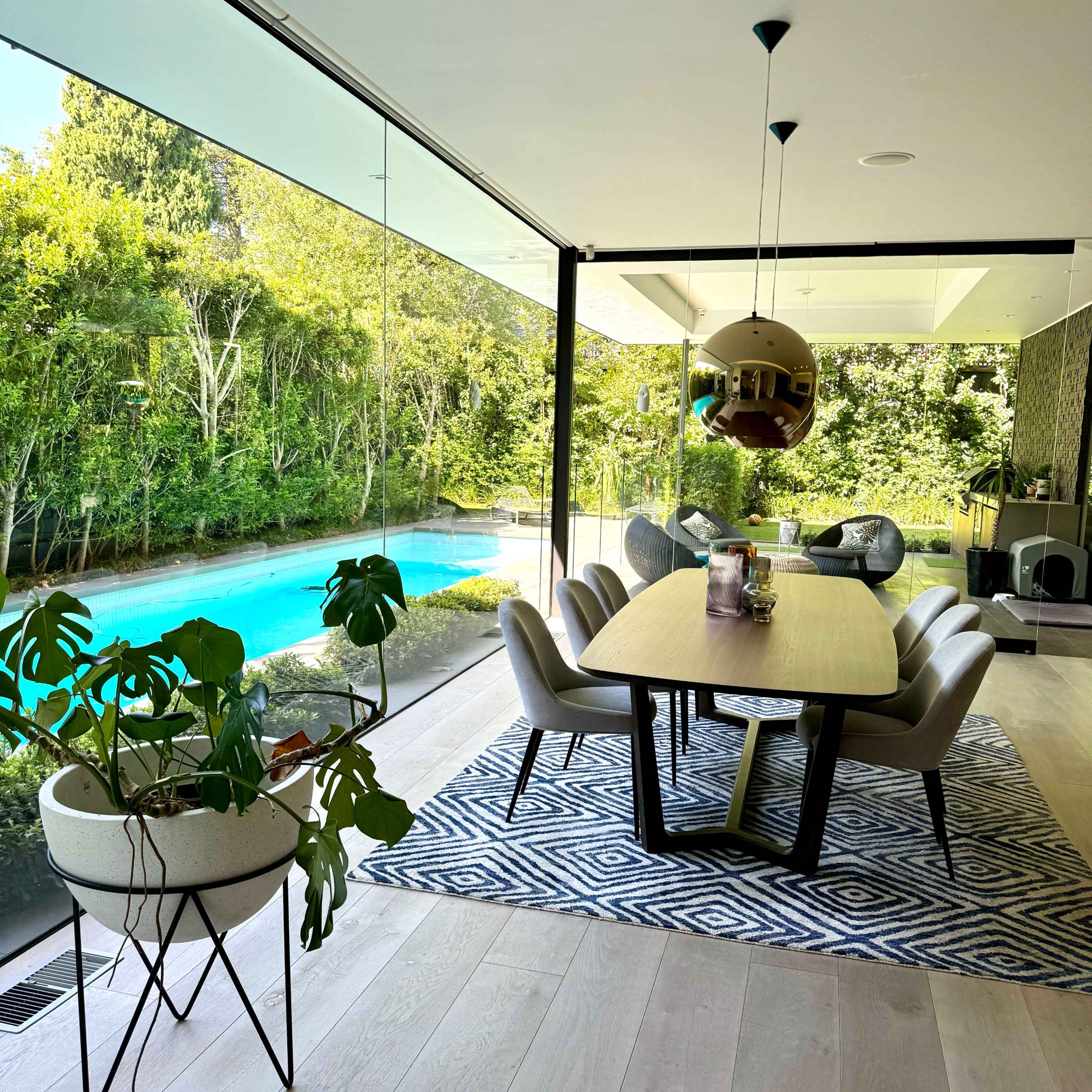 A modern dining area features a large table surrounded by chairs, with a view of a blue swimming pool and greenery through glass walls.