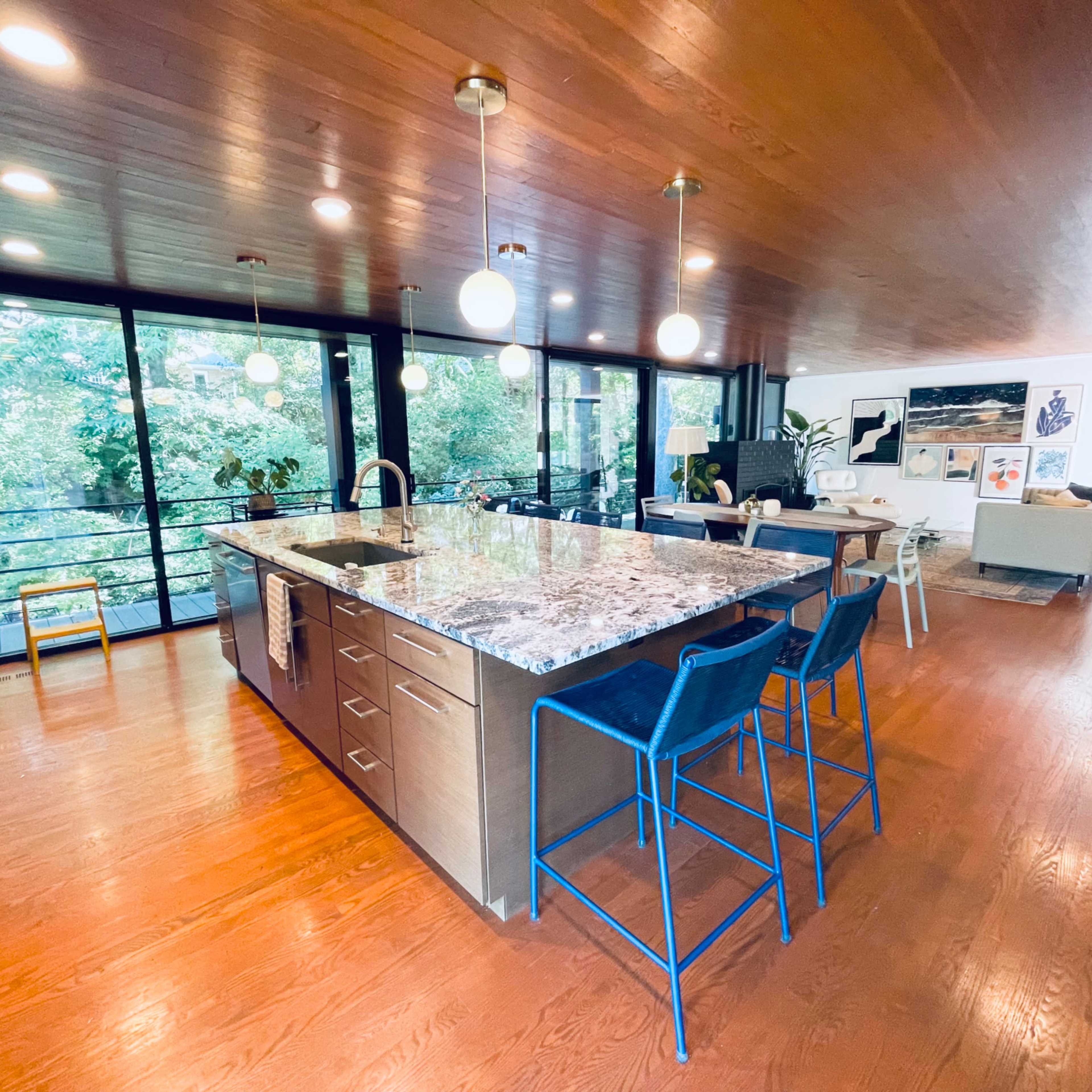 A spacious kitchen features a large granite island with blue bar stools, surrounded by large windows that provide a view of the greenery outside.