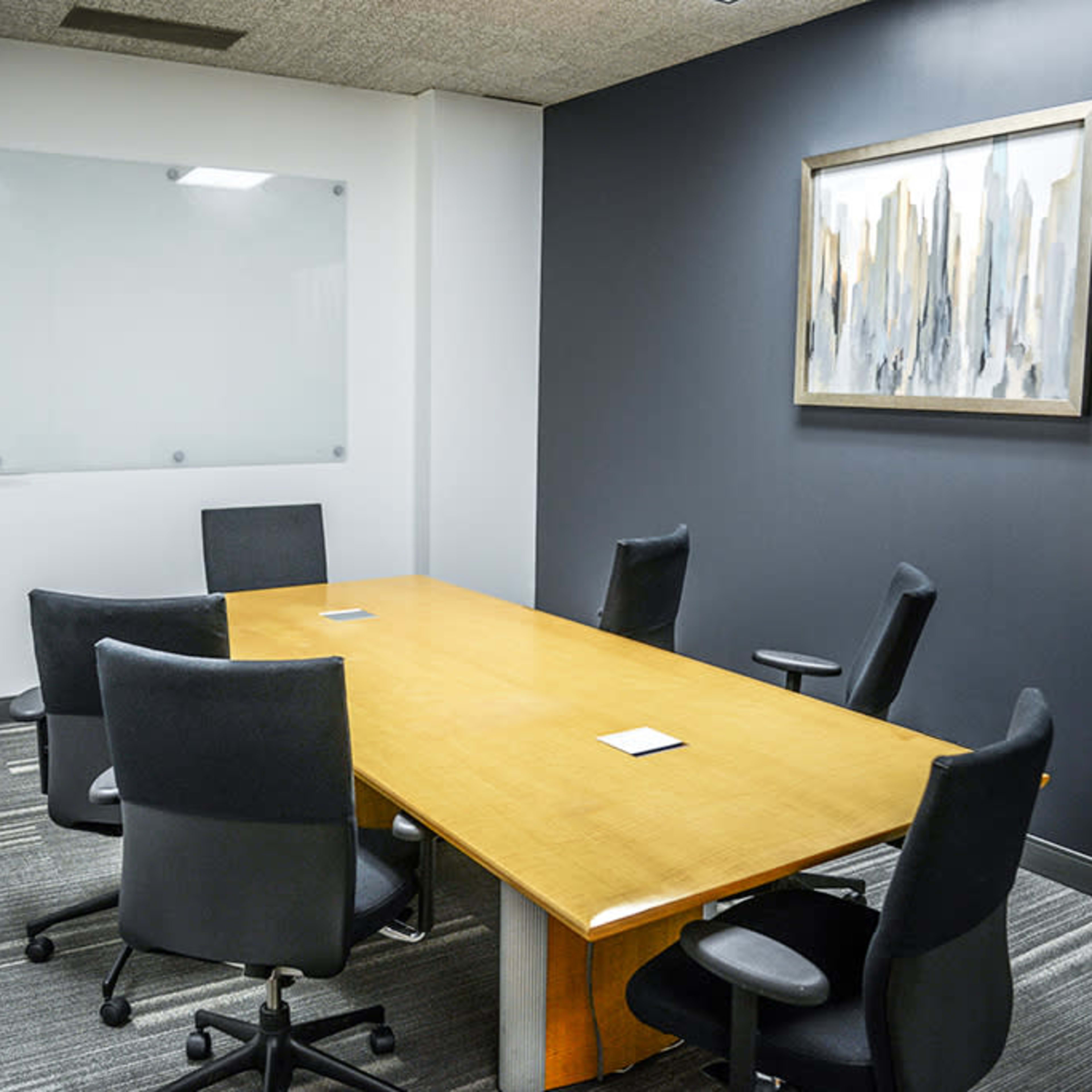 A conference room features a large wooden table surrounded by six black office chairs, with a whiteboard and a framed abstract painting on the walls.