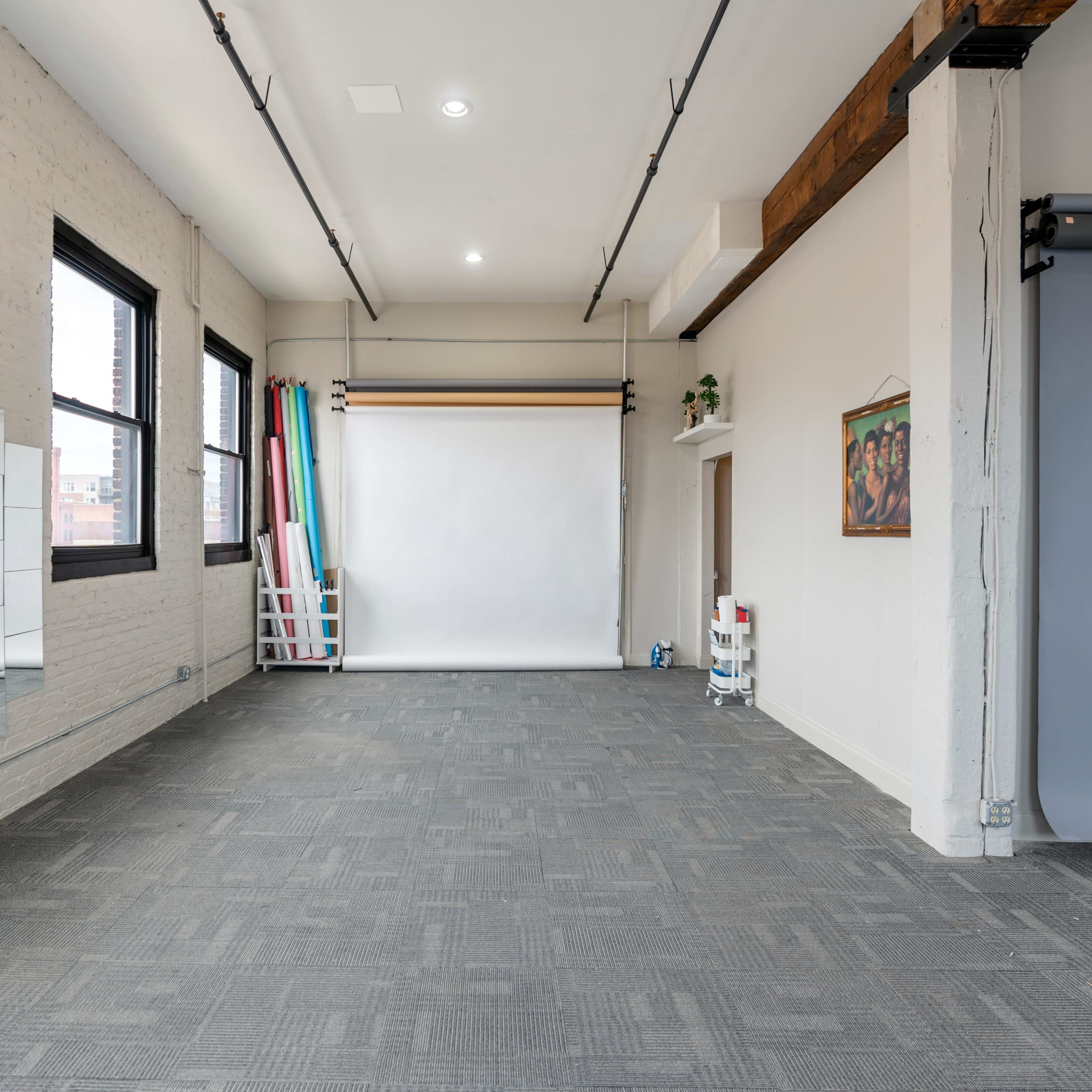 A spacious, empty studio with large windows, a white backdrop, and rolled backdrops in various colors against one wall.