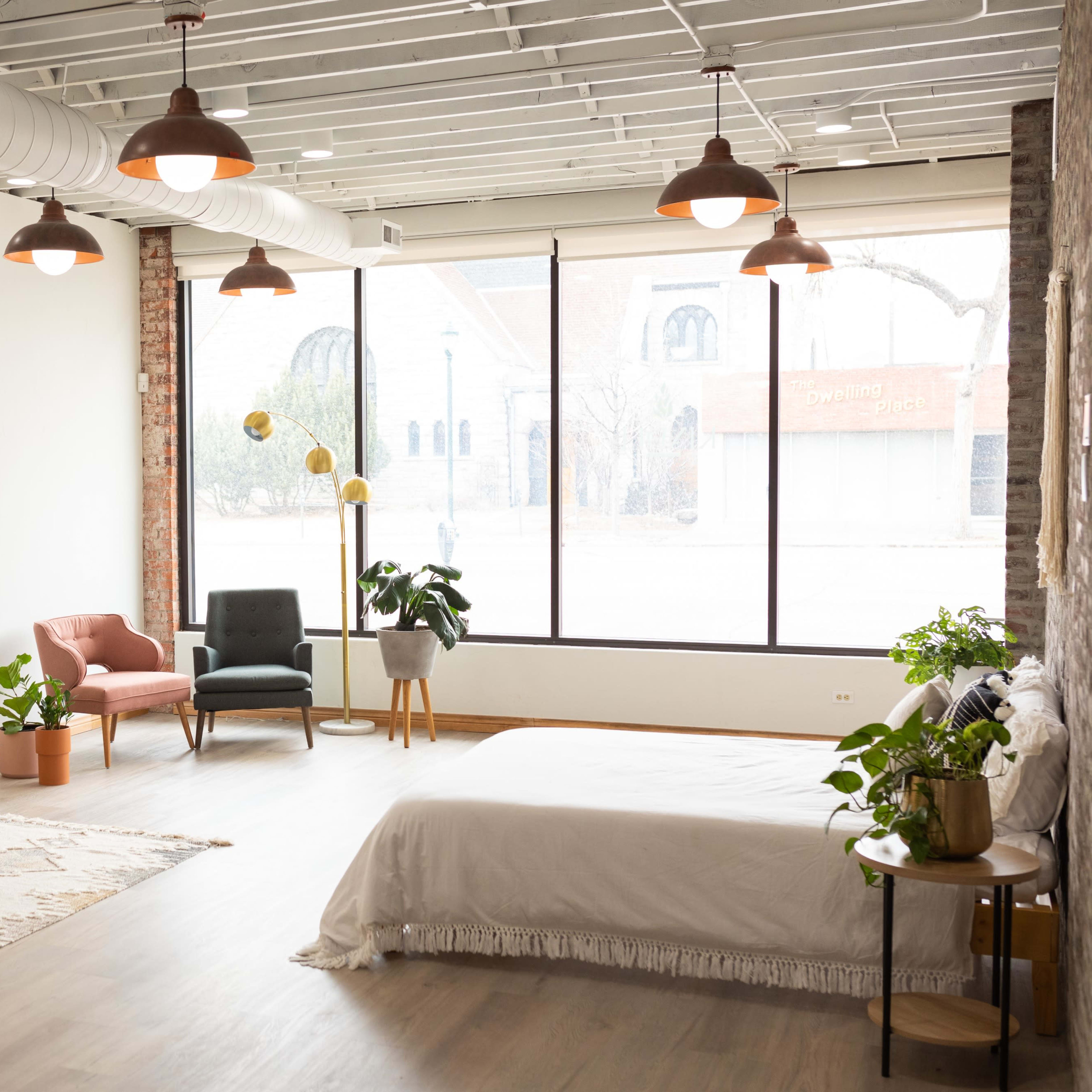A spacious, modern room featuring a bed, a sofa, two armchairs, and various indoor plants, with large windows allowing natural light to flood the space.