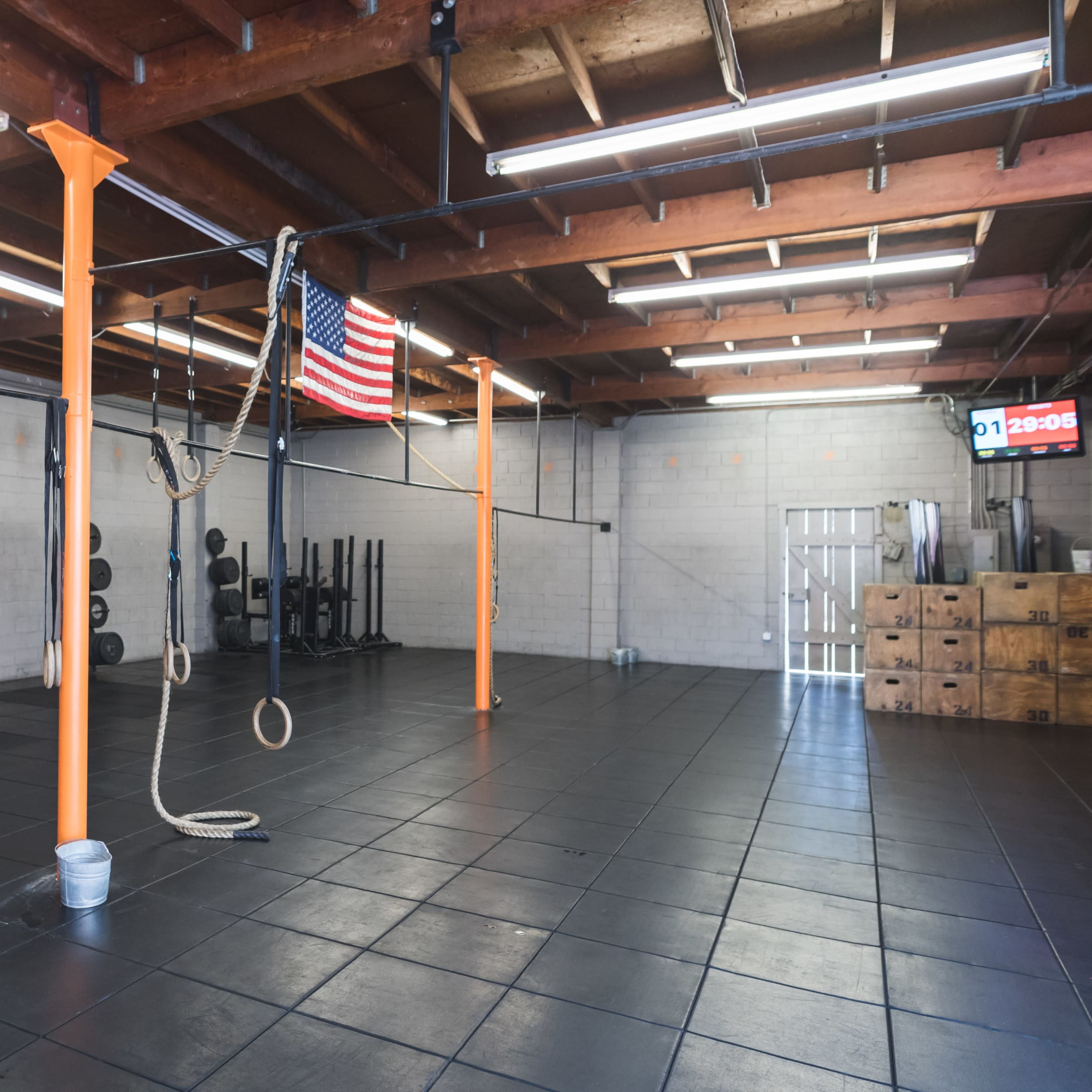A spacious gym interior with black rubber flooring, weight plates stacked against the walls, and gymnastic rings hanging from overhead bars, an American flag displayed, and a clock showing the time.