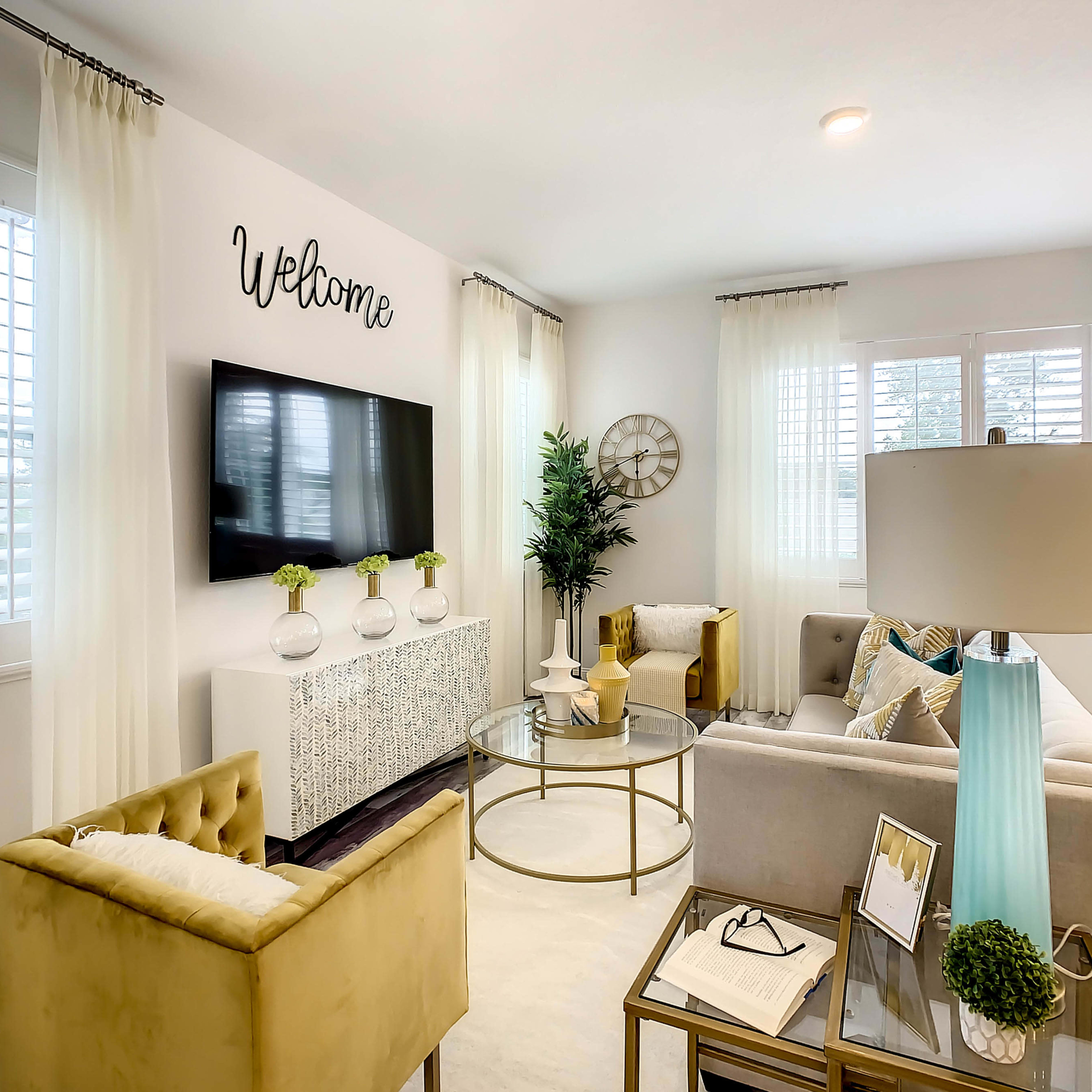 A modern living room with a light-colored sofa, a glass coffee table, and a television mounted on a white wall, decorated with a "Welcome" sign and plants.