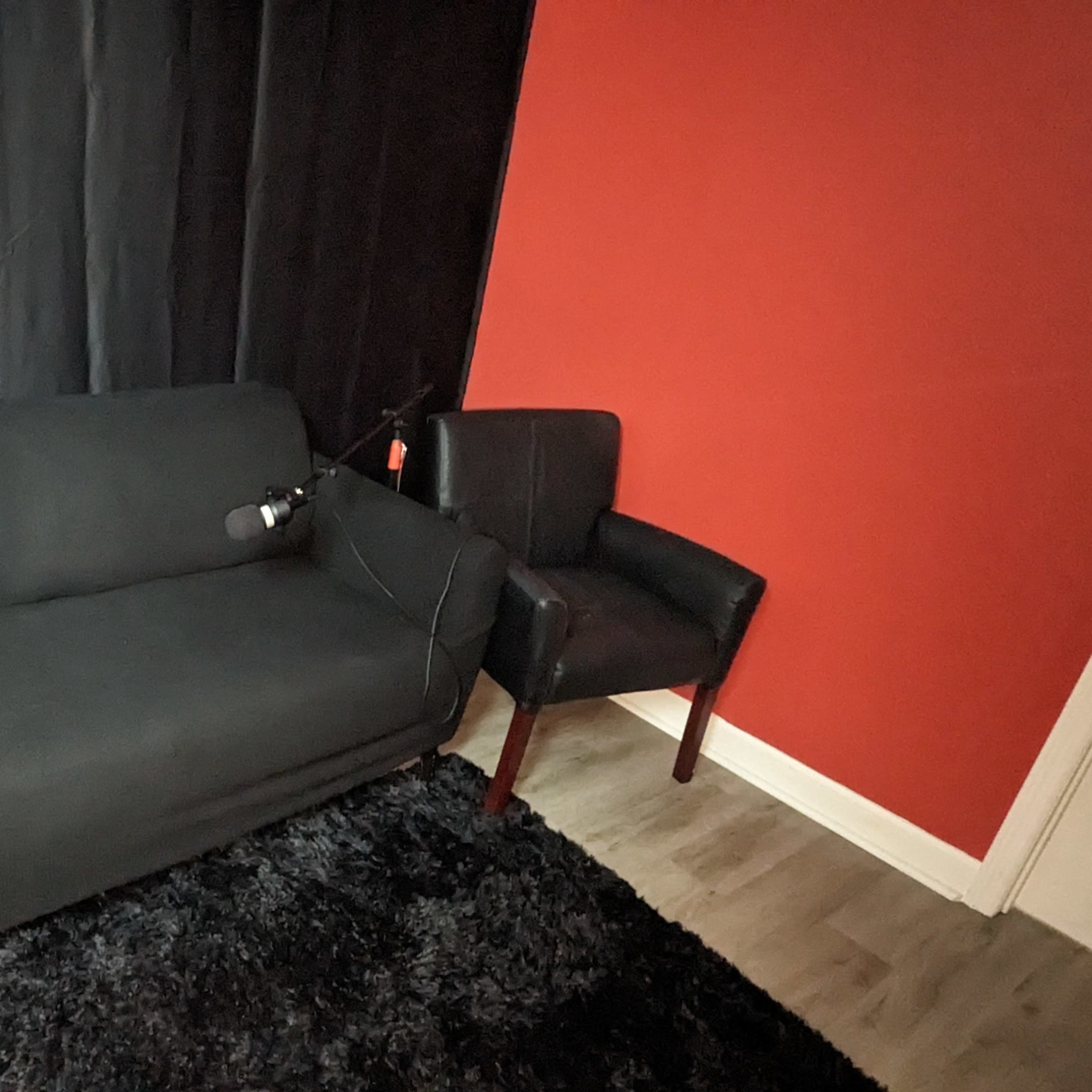 A black couch and a black armchair are positioned against a red wall, with microphones mounted on both chairs.