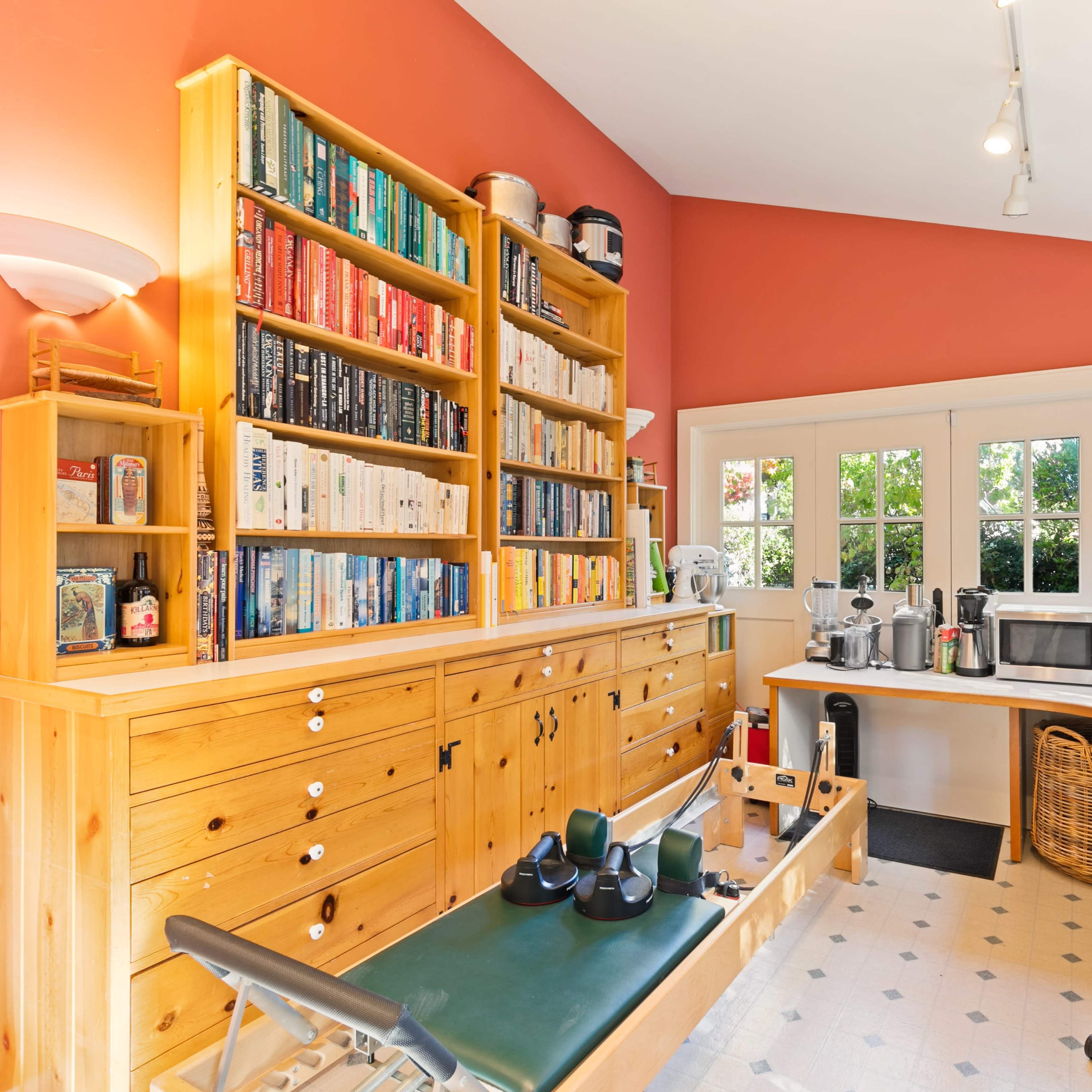 A brightly lit home office with wooden shelves filled with books, a desk with a computer, and a reformer Pilates machine.
