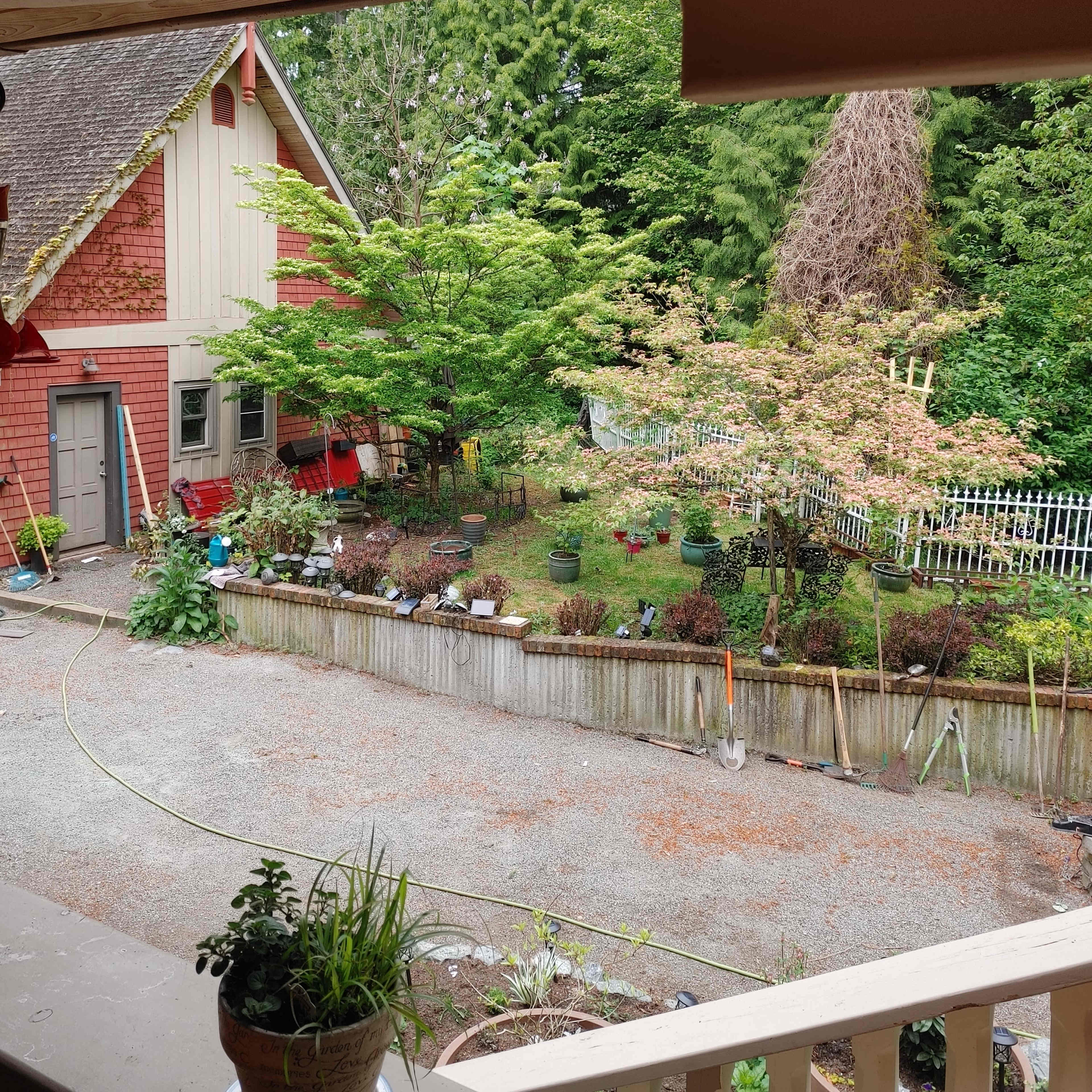 A view from a covered porch shows a landscaped garden with various plants and trees behind a quaint house.